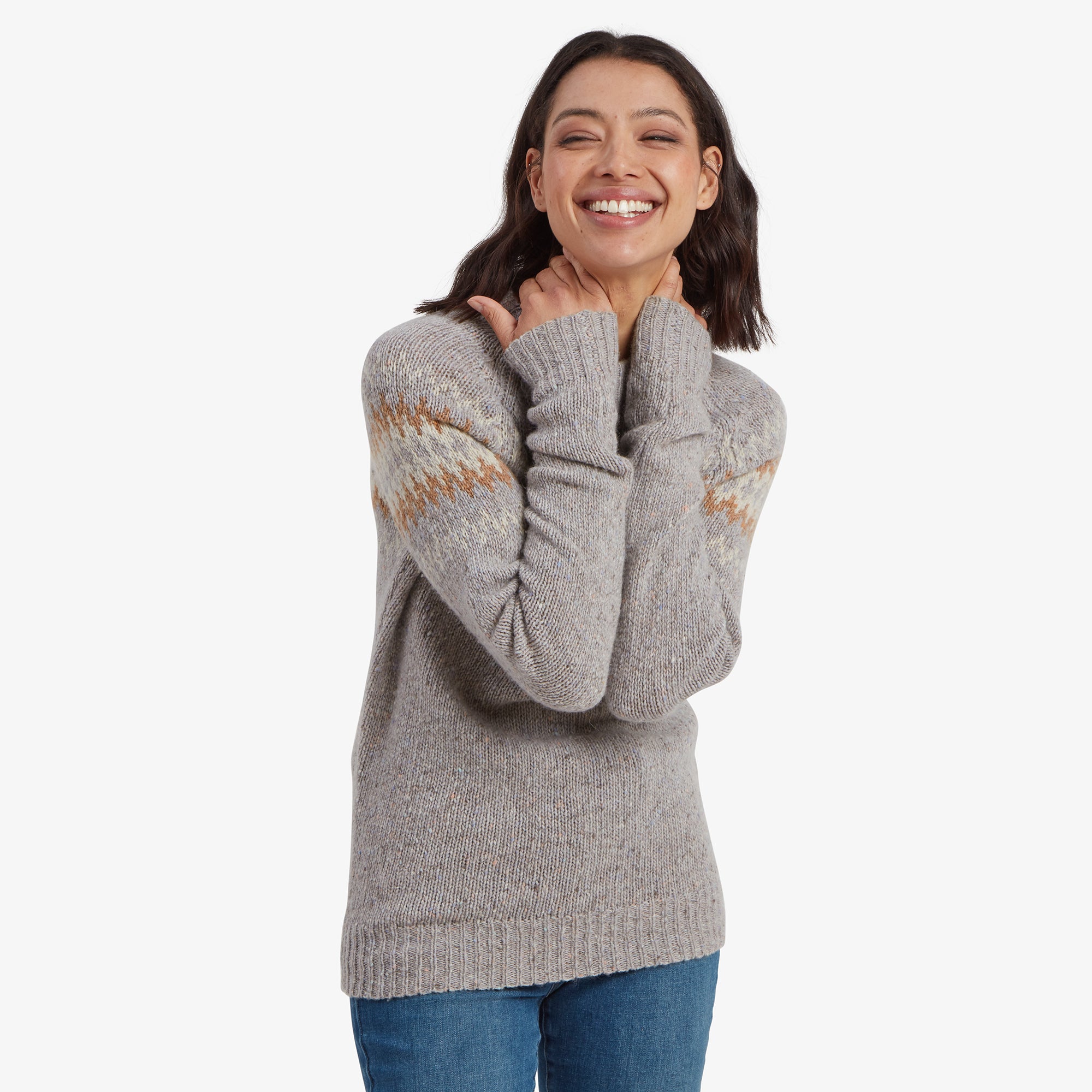 A woman smiles brightly, wearing a Sherpa Adventure Gear Dumji Crew Sweater in Grey with warm orange and cream zigzag patterns across the chest and upper sleeves. The high neck and textured knit detailing give it a warm and classic winter feel. Her relaxed, joyful expression and hands cuddling the collar enhance the cozy aesthetic.