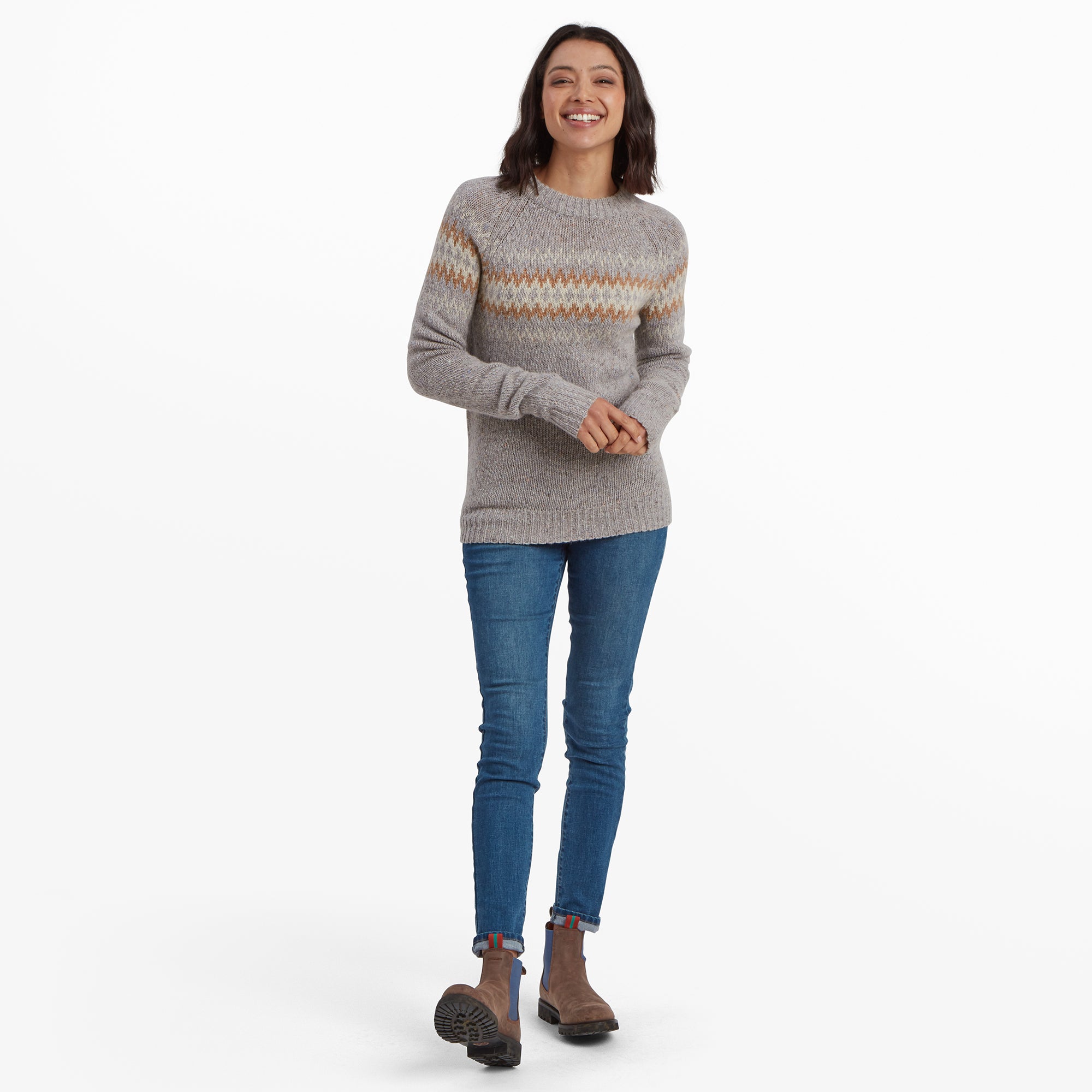 The same woman is shown standing in a full-length view, styled in blue skinny jeans and brown ankle boots. Her Sherpa Adventure Gear Dumji Crew Sweater in Grey’s soft texture and slightly relaxed fit are visible, along with the vibrant zigzag pattern. The outfit is casual yet stylish, perfect for cooler weather.