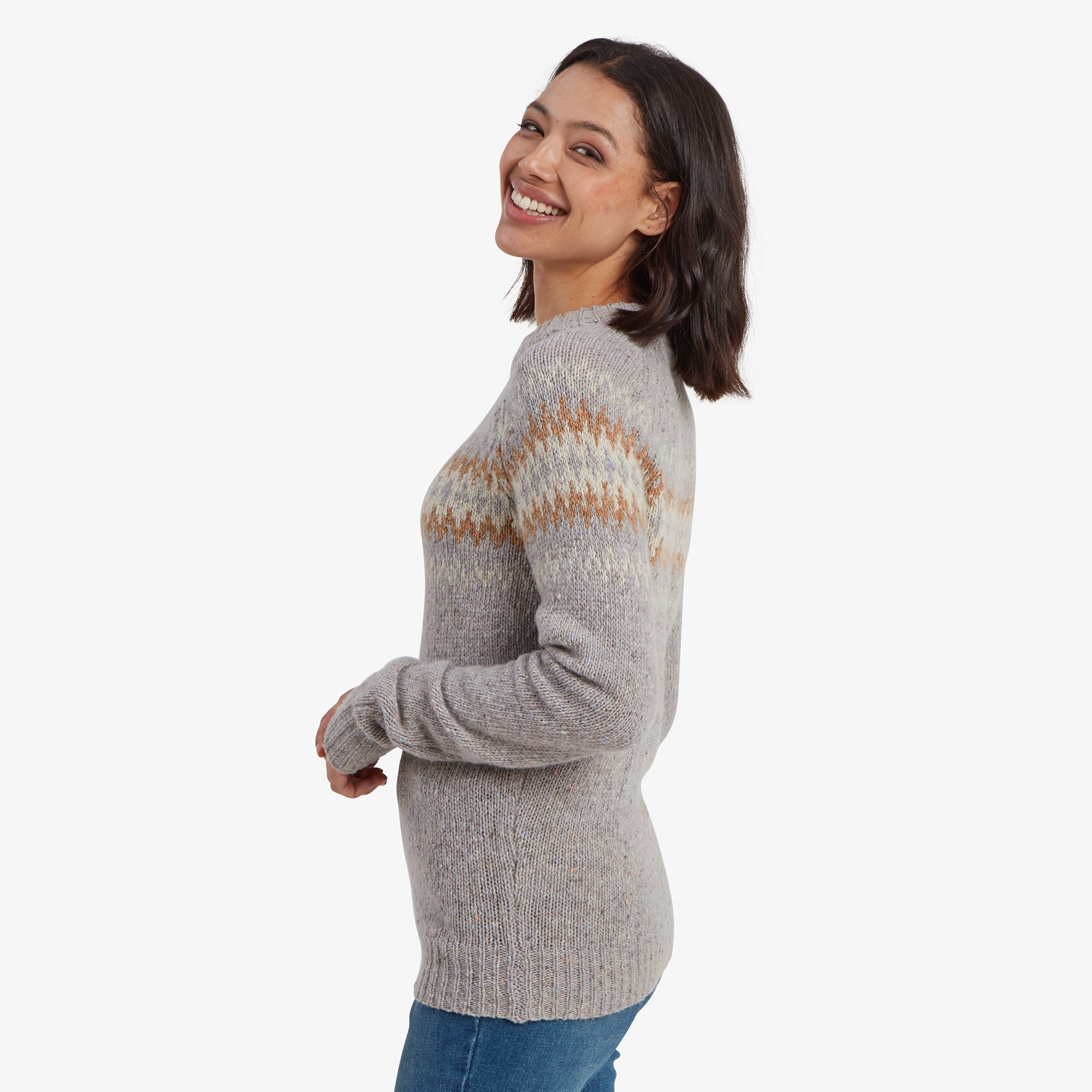 A side profile of the woman highlights the Sherpa Adventure Gear Dumji Crew Sweater in Grey’s fit and detailing. The zigzag pattern continues seamlessly across the arms, while the sweater’s ribbed hem and slightly elongated length provide a flattering silhouette. She looks over her shoulder with a cheerful expression.