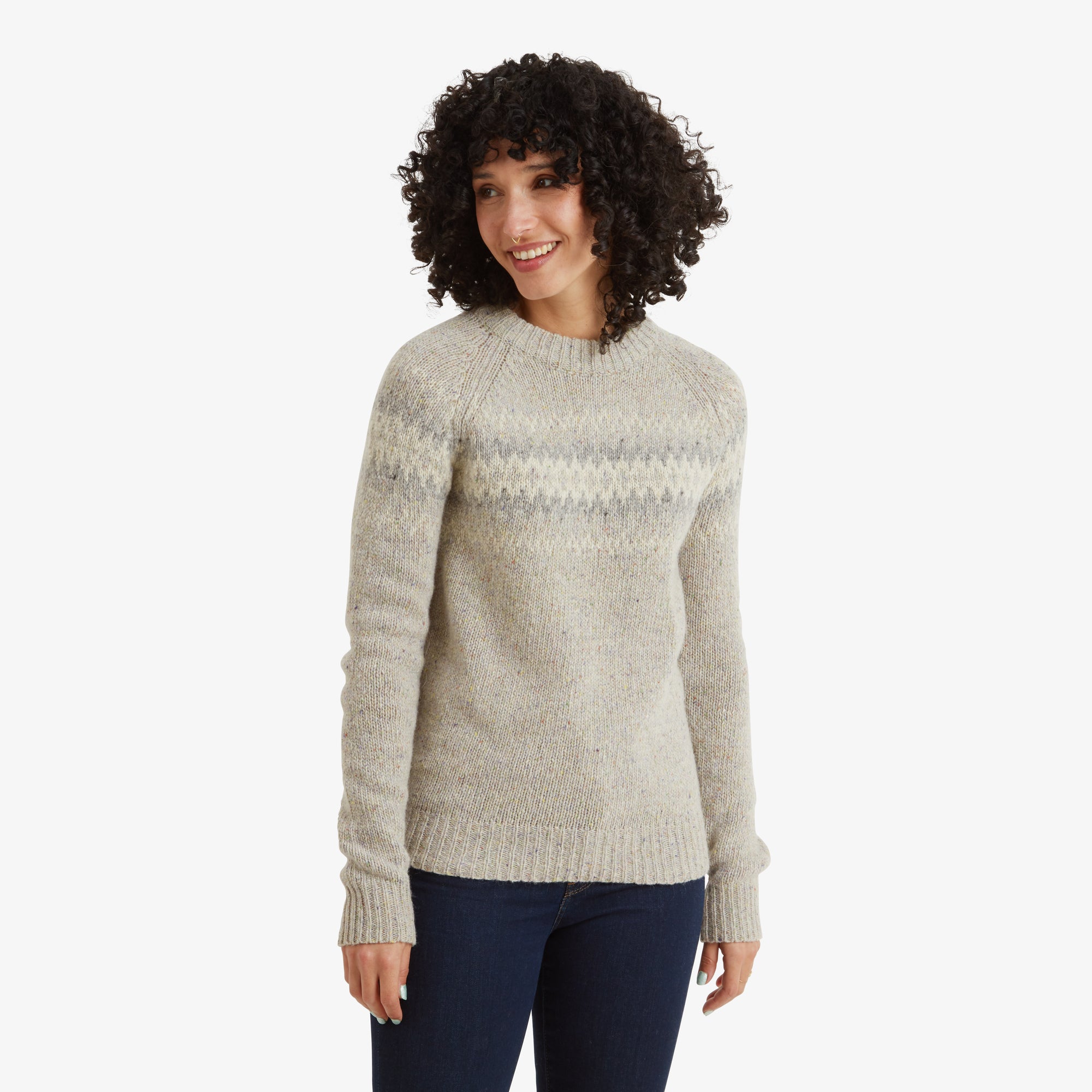 A woman with curly dark hair is wearing a Sherpa Adventure Gear Dumji Crew Sweater in White featuring a subtle geometric pattern in gray across the chest and upper arms. The sweater has a classic crew neck and ribbed cuffs. She is smiling and standing casually, paired with dark jeans.