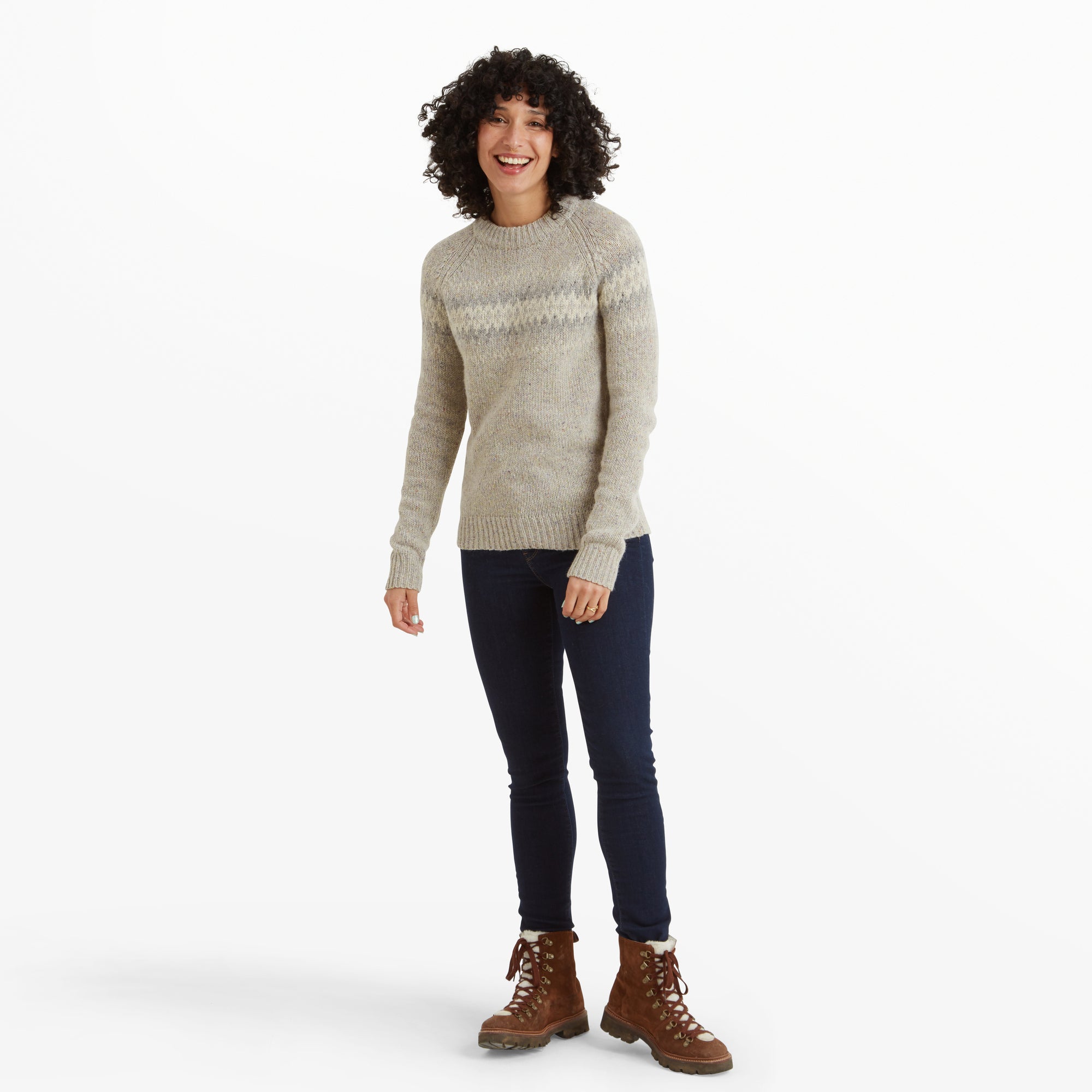 The woman showcases the Sherpa Adventure Gear Dumji Crew Sweater in White with a relaxed stance, smiling broadly. She pairs the light beige and gray-patterned sweater with dark skinny jeans and brown lace-up hiking boots, emphasising a casual and outdoorsy look.