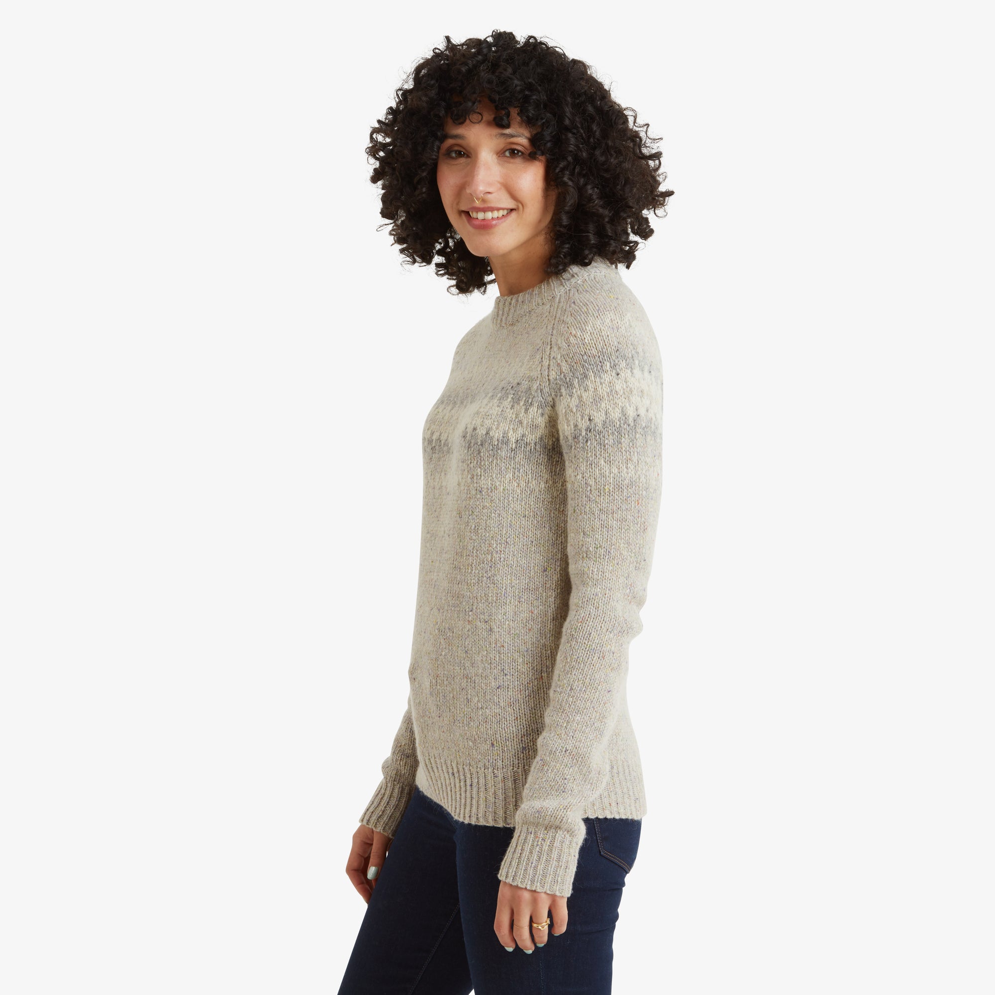 The same Sherpa Adventure Gear Dumji Crew Sweater in White is shown in profile, highlighting the side fit and detailing of the pattern. The geometric design extends seamlessly around the sweater, and the ribbed hem is visible. The woman is smiling subtly, facing slightly forward.