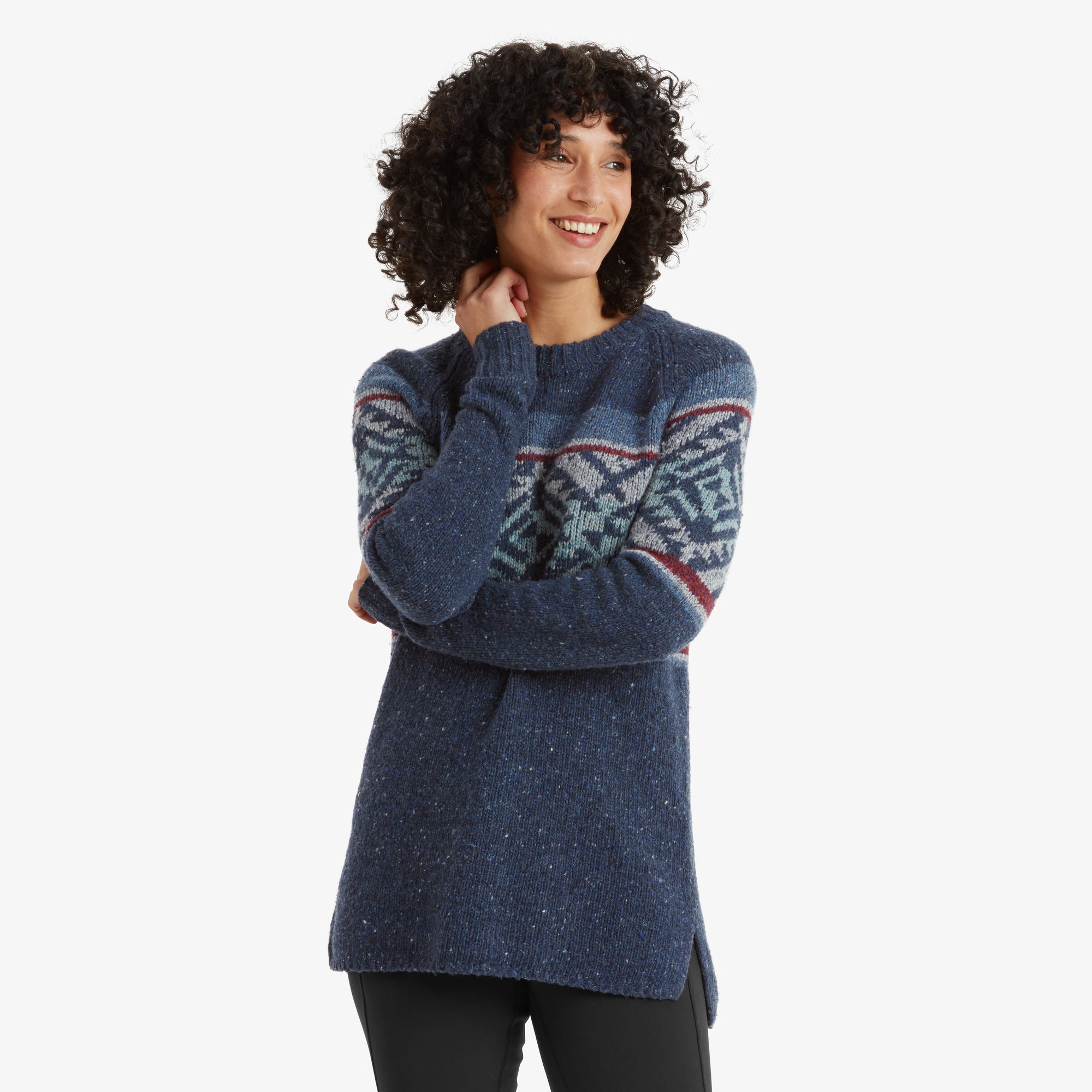 A close-up of a woman wearing a Sherpa Adventure Gear Dumji Long Sweater in Blue with a geometric Nordic-inspired pattern in teal, white, and red on the chest and sleeves. She is smiling with her head slightly tilted and arms crossed, showing the texture and details of the sweater fabric.