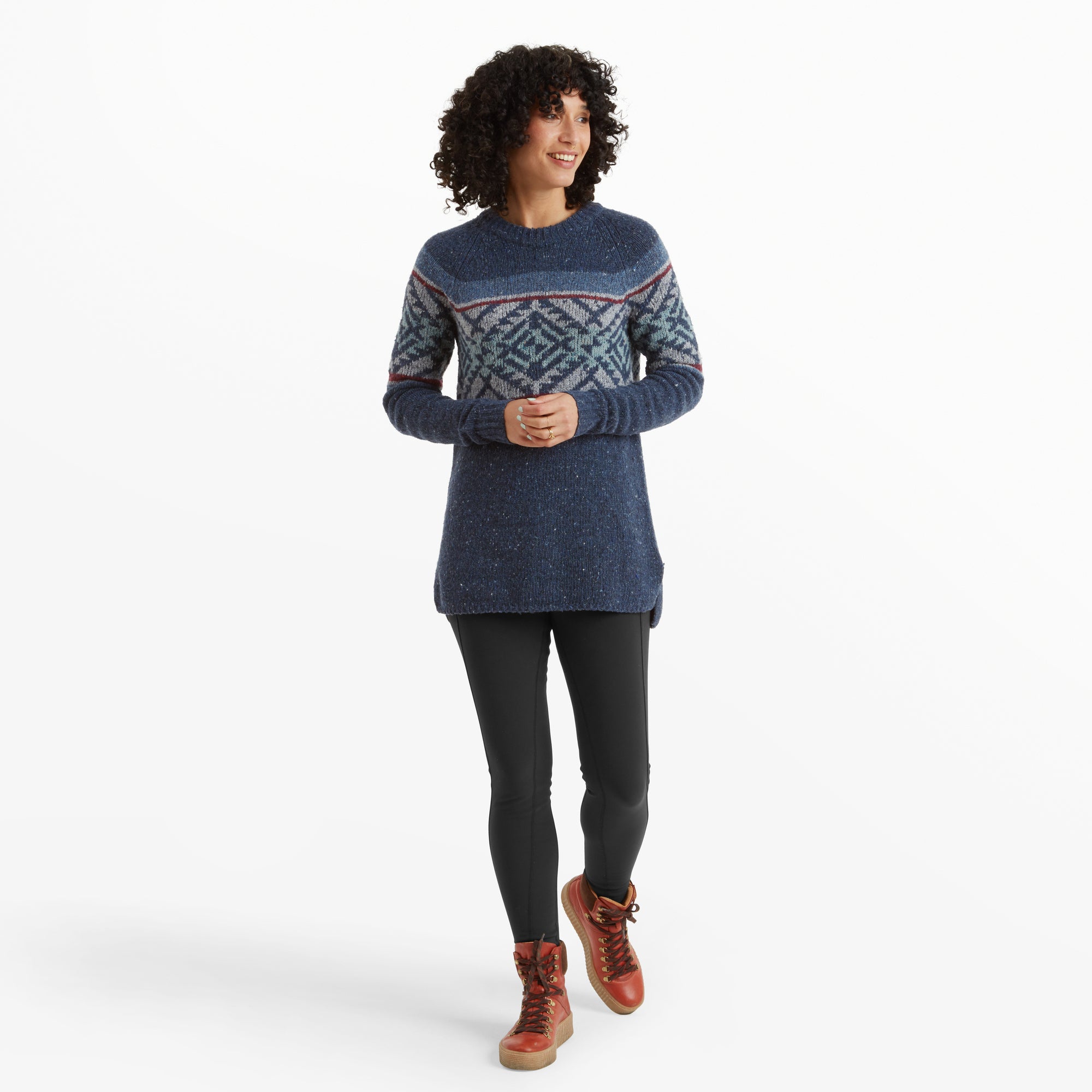 A full-body image of the same woman wearing the Sherpa Adventure Gear Dumji Long Sweater in Blue paired with black leggings and red hiking boots. She stands confidently with her hands clasped in front of her, showcasing the relaxed fit and slightly longer back hem of the sweater.