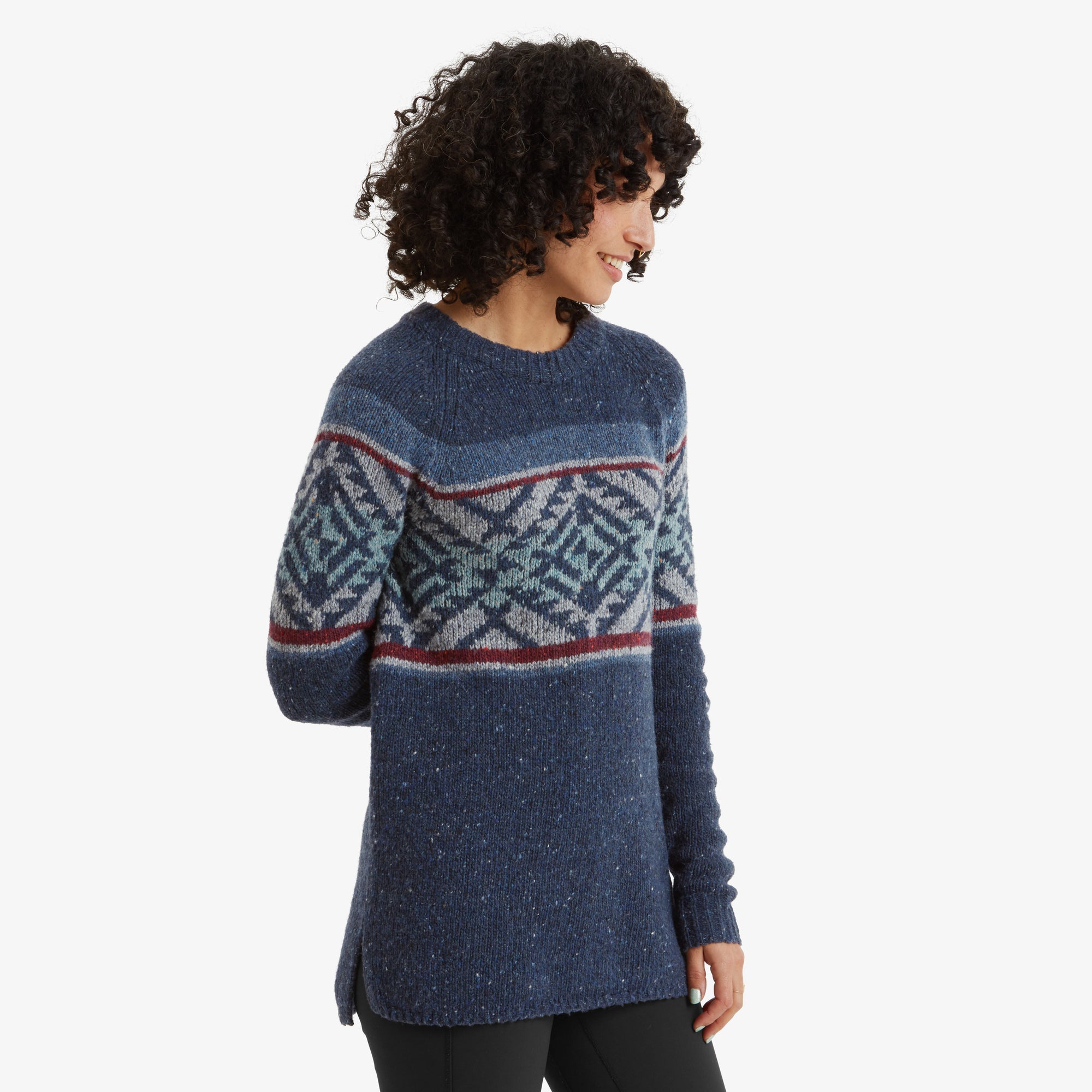 A side profile of the woman wearing the Sherpa Adventure Gear Dumji Long Sweater in Blue. She looks down with a soft smile, her curly hair framing her face. The geometric pattern wraps around the chest and arms, and the side slit detail at the hem of the sweater is visible.