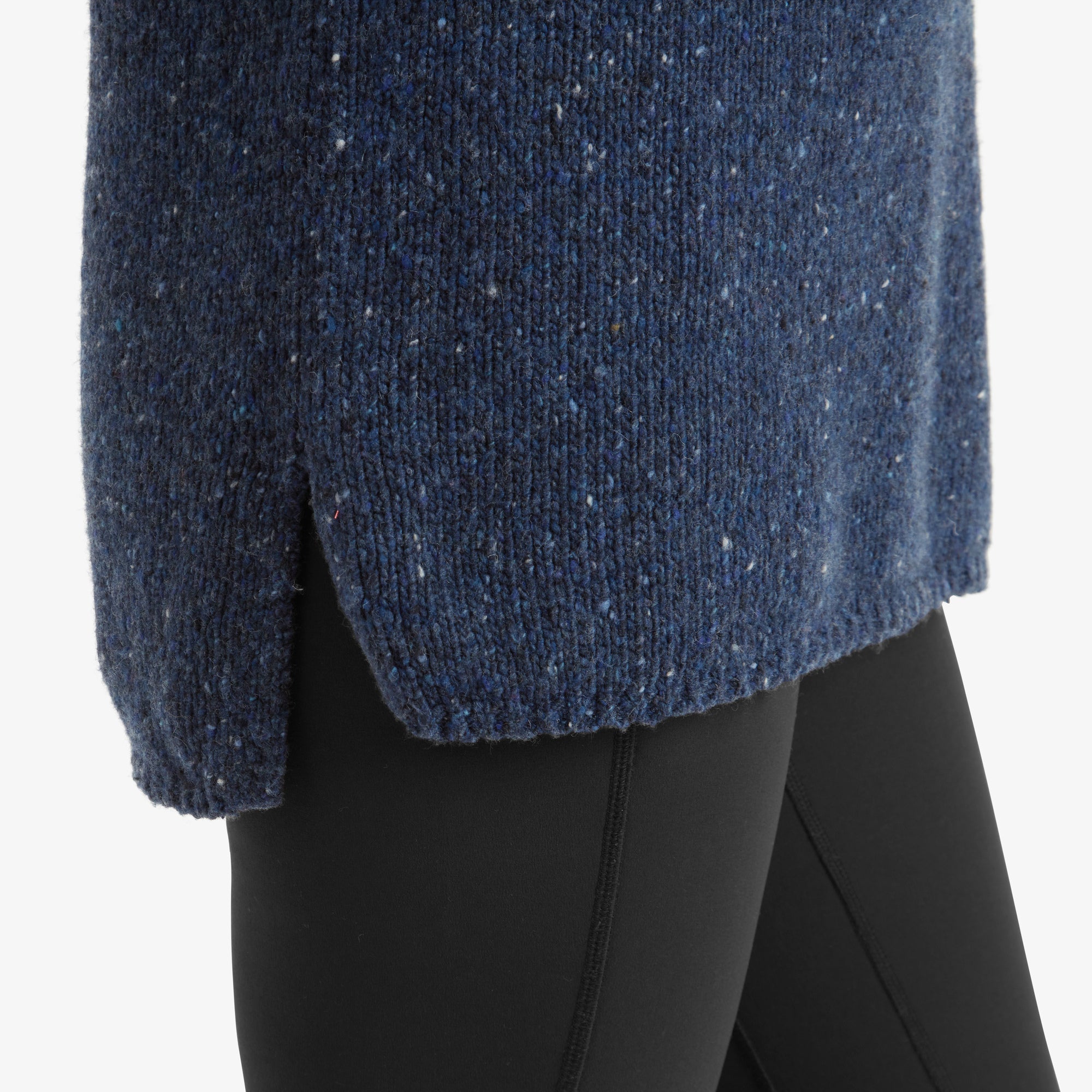 A close-up of the hemline of the Sherpa Adventure Gear Dumji Long Sweater in Blue, focusing on the side slit detail and textured knit. The sweater's rich blue fabric features subtle speckled flecks, paired with black leggings that emphasise the casual and comfortable fit.