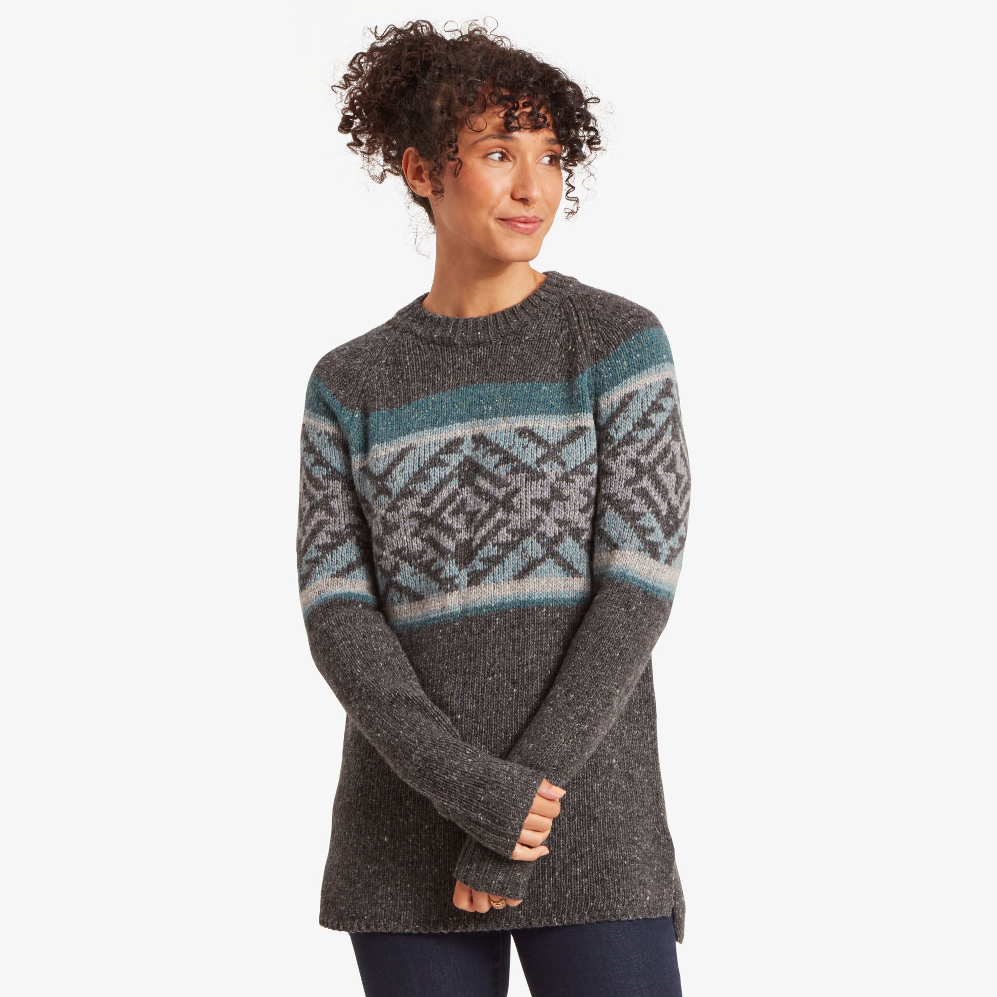 A model wearing a Sherpa Adventure Gear Dumji Long Sweater in Grey with a patterned yoke in blue and grey geometric designs. The sweater features a round neckline and long sleeves. The model's arms are slightly crossed in front, and she smiles softly, creating a cozy and stylish look.