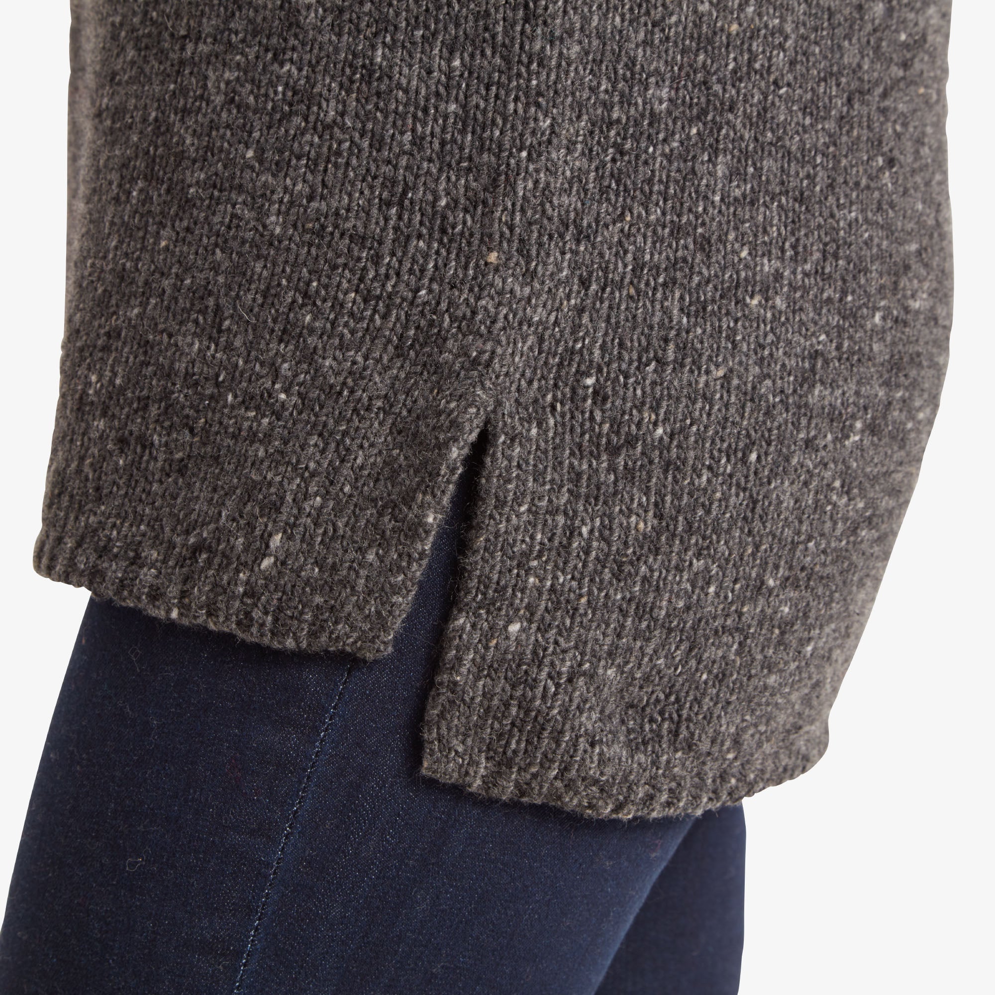 A close-up of the Sherpa Adventure Gear Dumji Long Sweater in Grey's hem on one side, highlighting the small side slit for added comfort and movement. The textured knit of the fabric is evident against dark denim jeans.