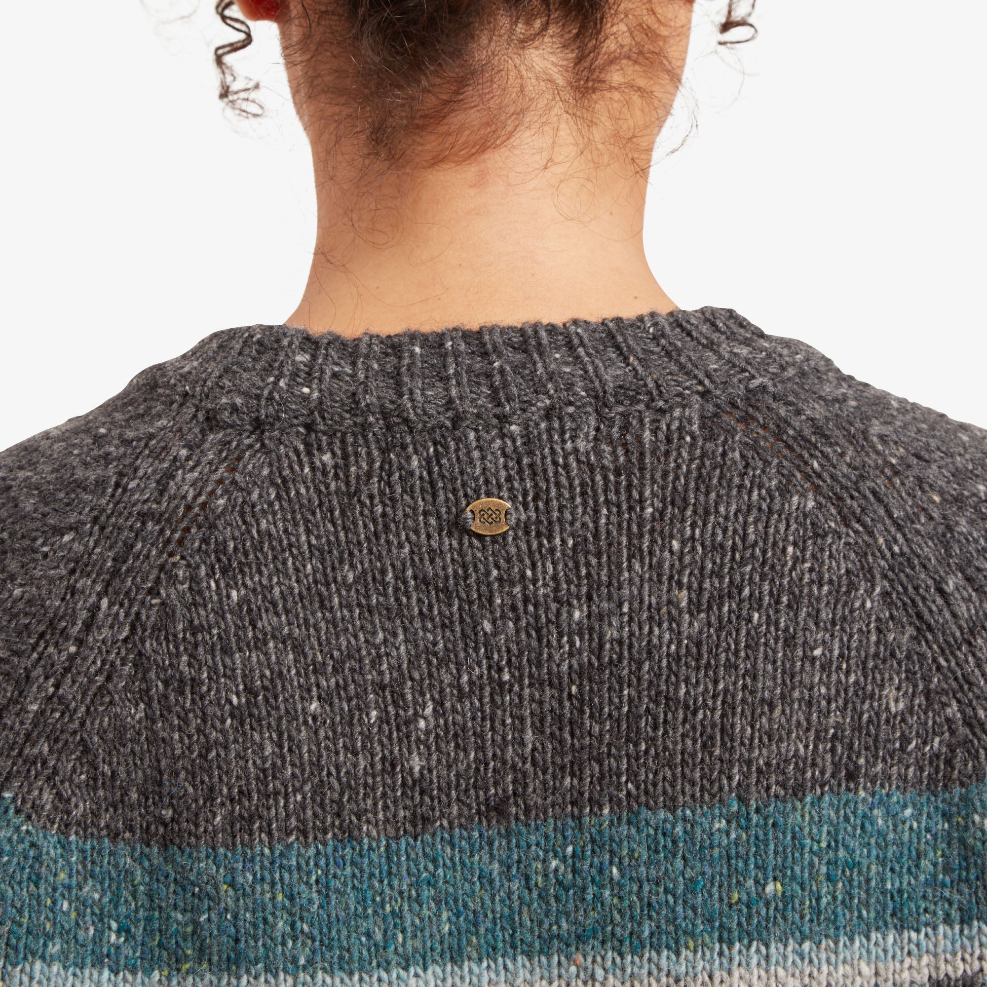 A zoomed-in view of the brass Sherpa logo on the back of the Sherpa Adventure Gear Dumji Long Sweater in Grey, situated at the center of the yoke. The intricate geometric pattern in the yoke adds depth to the design.