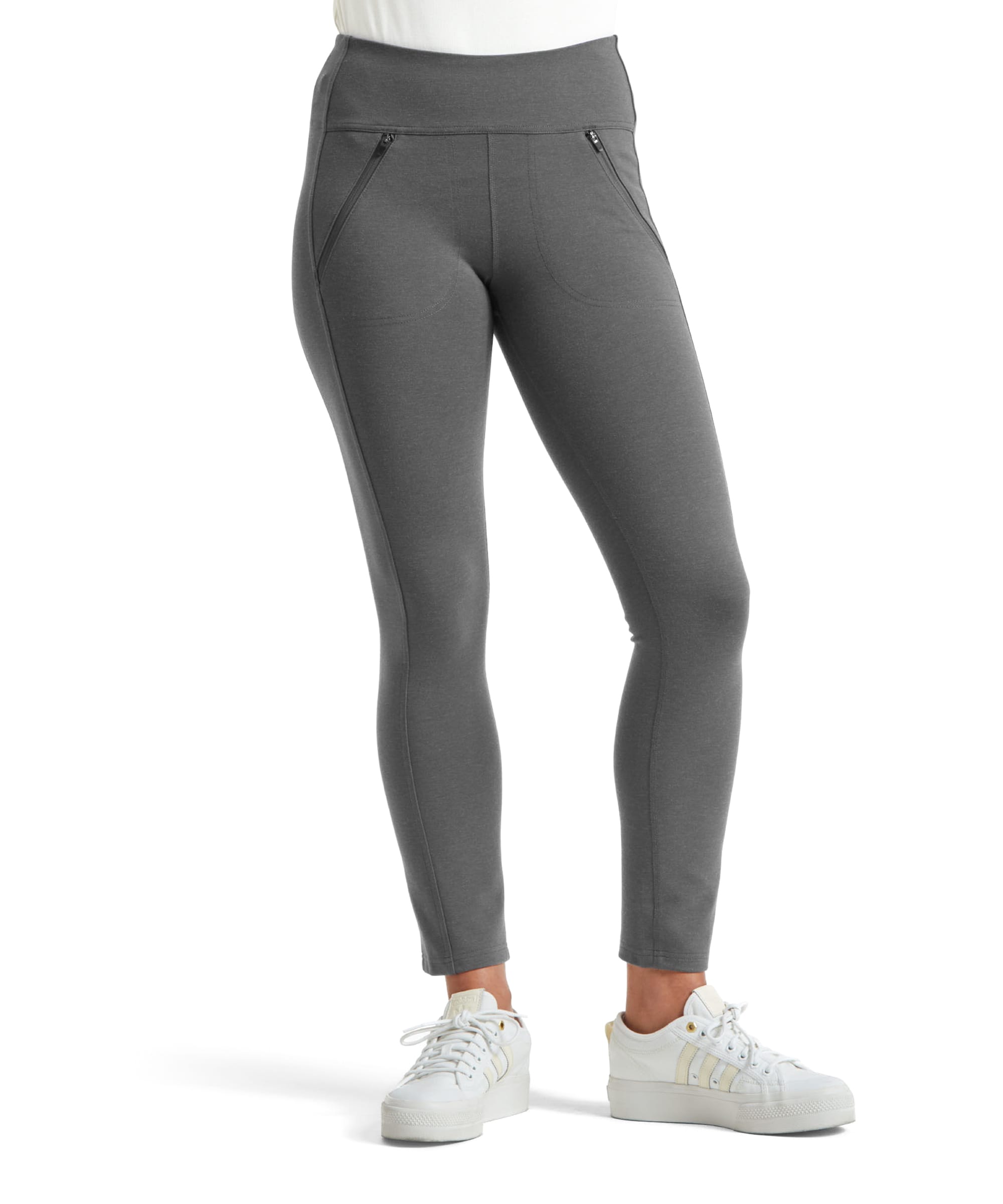 A close-up of the Sherpa Adventure Gear Hapta Knit trousers in Grey. The image focuses on the lower half of a female model, showcasing the slim-fit leggings. The trousers have a high-rise, wide waistband for comfort and two visible front zippered pockets with sleek stitching. The model pairs the trousers with white trainers, highlighting a casual yet polished look. The trousers end at ankle length, providing a snug and tailored appearance.