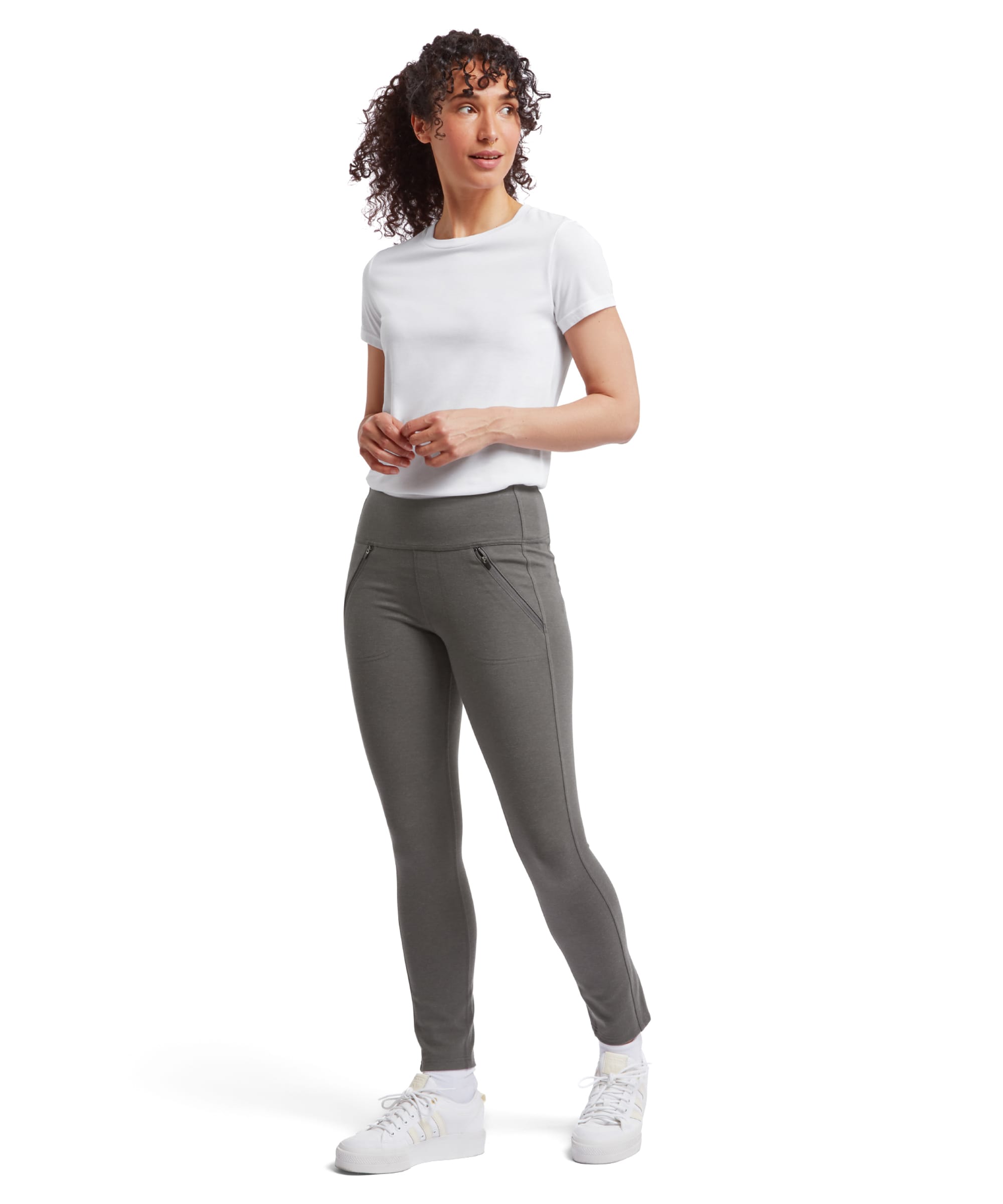 A full-body shot of a female model wearing the Sherpa Adventure Gear Hapta Knit trousers in Grey paired with a plain white short-sleeve T-shirt. The model is standing at an angle, smiling softly while looking slightly to the side. Her curly hair cascades down her shoulders. The trousers are form-fitting, accentuating the streamlined silhouette, with zippered front pockets and an ankle-length cut. She wears white platform trainers, adding a casual, sporty vibe to the outfit.