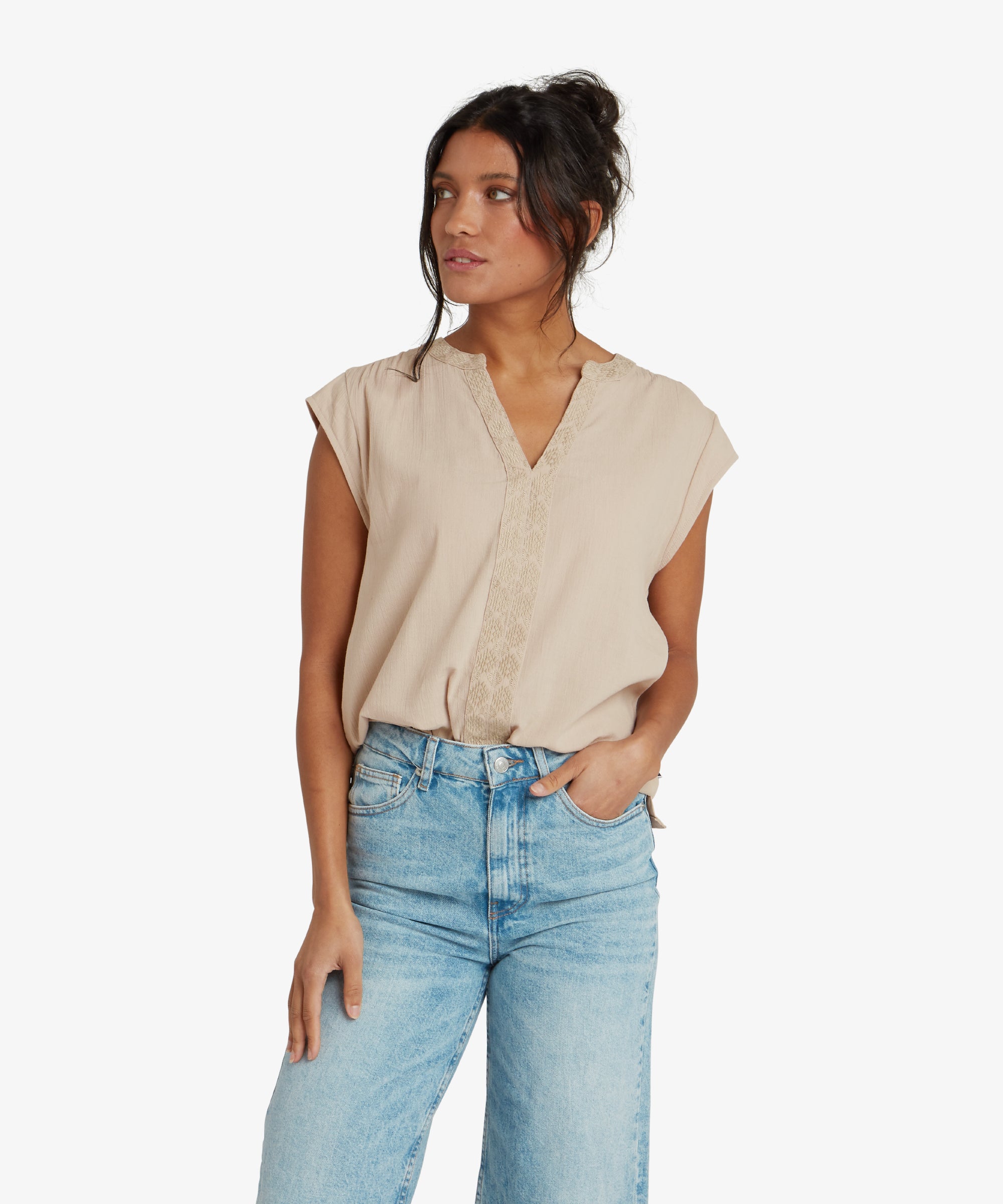 A woman is wearing a Sherpa Adventure Gear Hara Cap Sleeve Top in Beige with a relaxed fit. The top features subtle embroidered details running vertically along the neckline. She is styled casually with blue jeans, smiling confidently toward the camera.