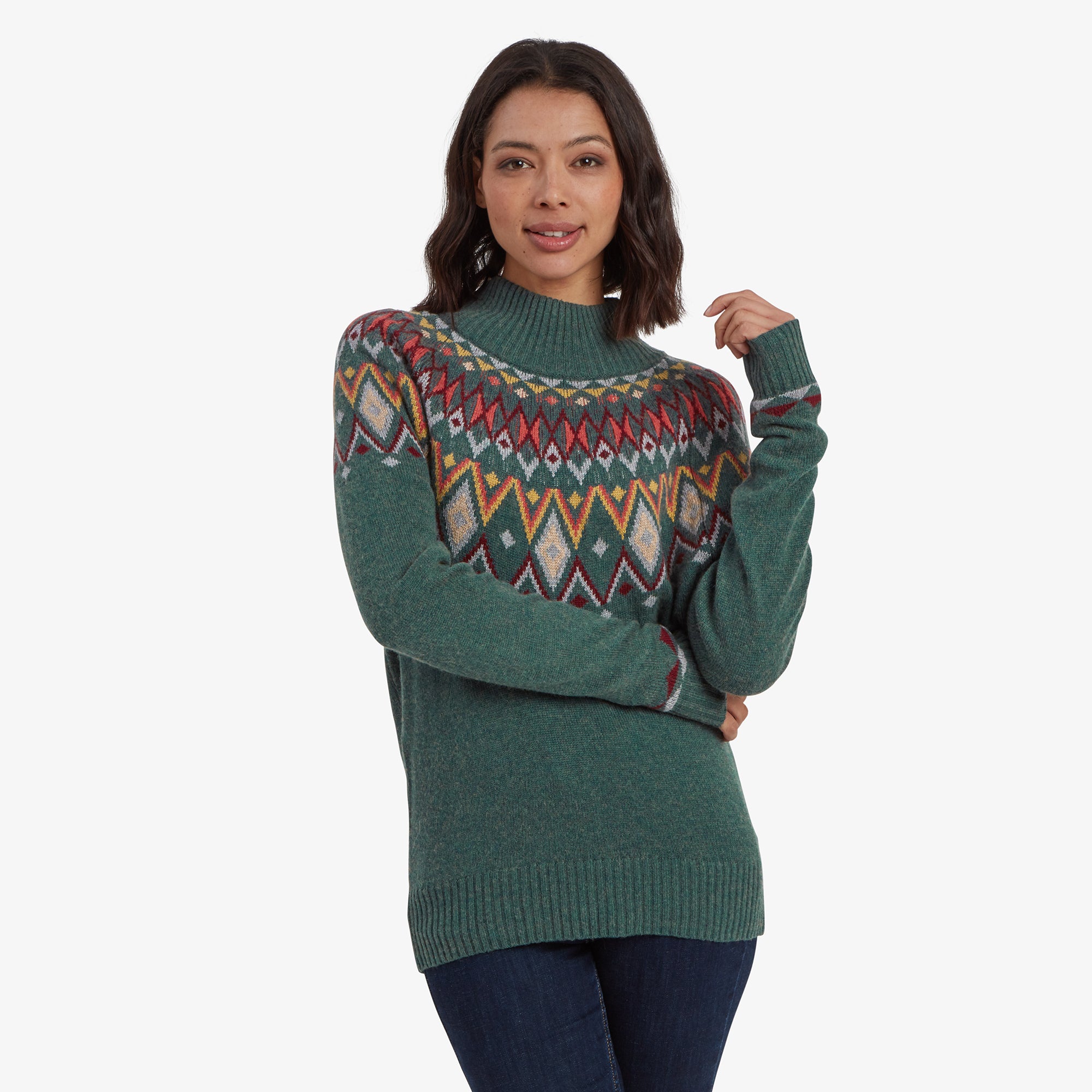 A woman wearing a Sherpa Adventure Gear Indu Eco Mock Neck in Green featuring a vibrant Fair Isle pattern across the chest and shoulders in red, yellow, and gray tones. She is standing with one hand near her face, smiling softly, and paired with dark denim jeans.