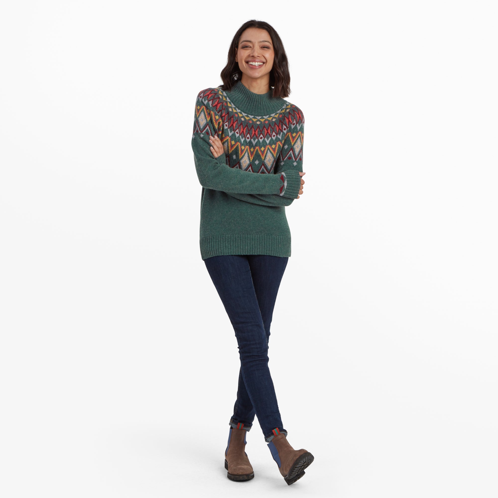 A woman smiling broadly while modeling the Sherpa Adventure Gear Indu Eco Mock Neck in Green with a colorful geometric Fair Isle design. She is standing with her arms folded, wearing dark denim jeans and brown ankle boots.