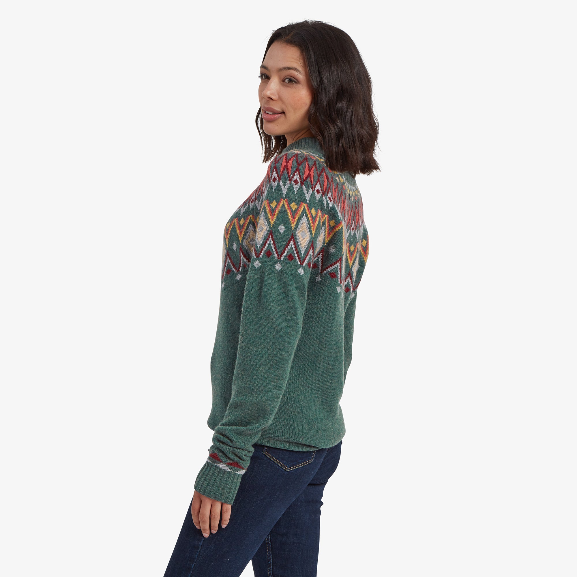 The woman showcasing the Sherpa Adventure Gear Indu Eco Mock Neck in Green's side profile. The bold Fair Isle design continues around the back, paired with dark jeans, and her hair styled in loose waves.