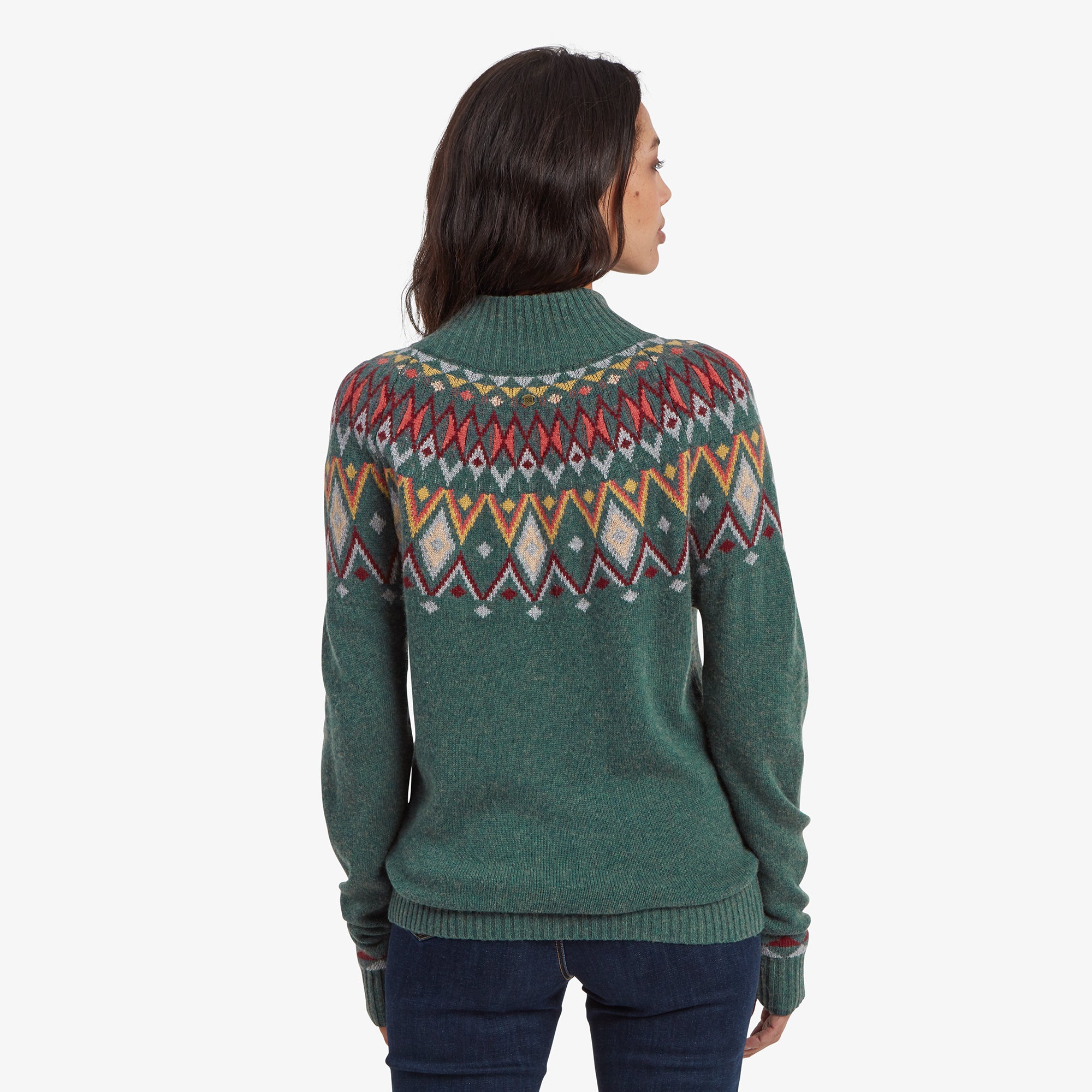 A rear view of the Sherpa Adventure Gear Indu Eco Mock Neck in Green with its colorful geometric pattern wrapping around the shoulders. The sweater fits loosely, and the model's hair is styled in a shoulder-length bob.