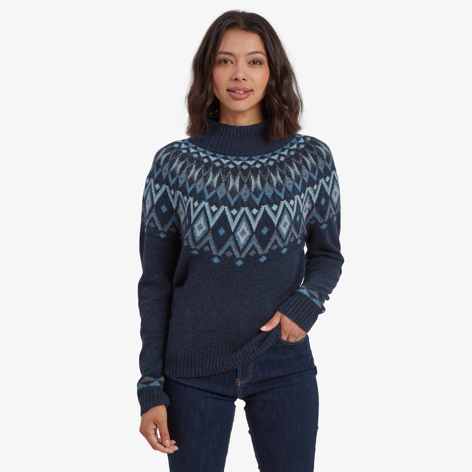 A woman with shoulder-length dark hair is standing against a white background, wearing a Sherpa Adventure Gear Indu Eco Mock Neck Sweater in Blue with a Nordic-inspired geometric pattern in shades of blue and white across the chest and upper arms. She has one hand casually tucked into her jeans pocket and is smiling softly.