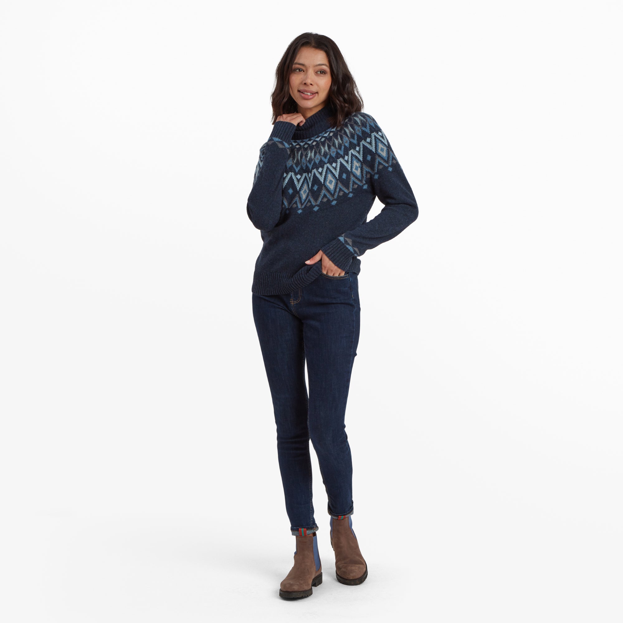 The same woman is posing with a relaxed stance, slightly tilting her head and placing one hand on her cheek while the other is in her pocket. She is wearing the Sherpa Adventure Gear Indu Eco Mock Neck Sweater in Blue, dark skinny jeans and brown ankle boots, completing a cozy winter outfit.