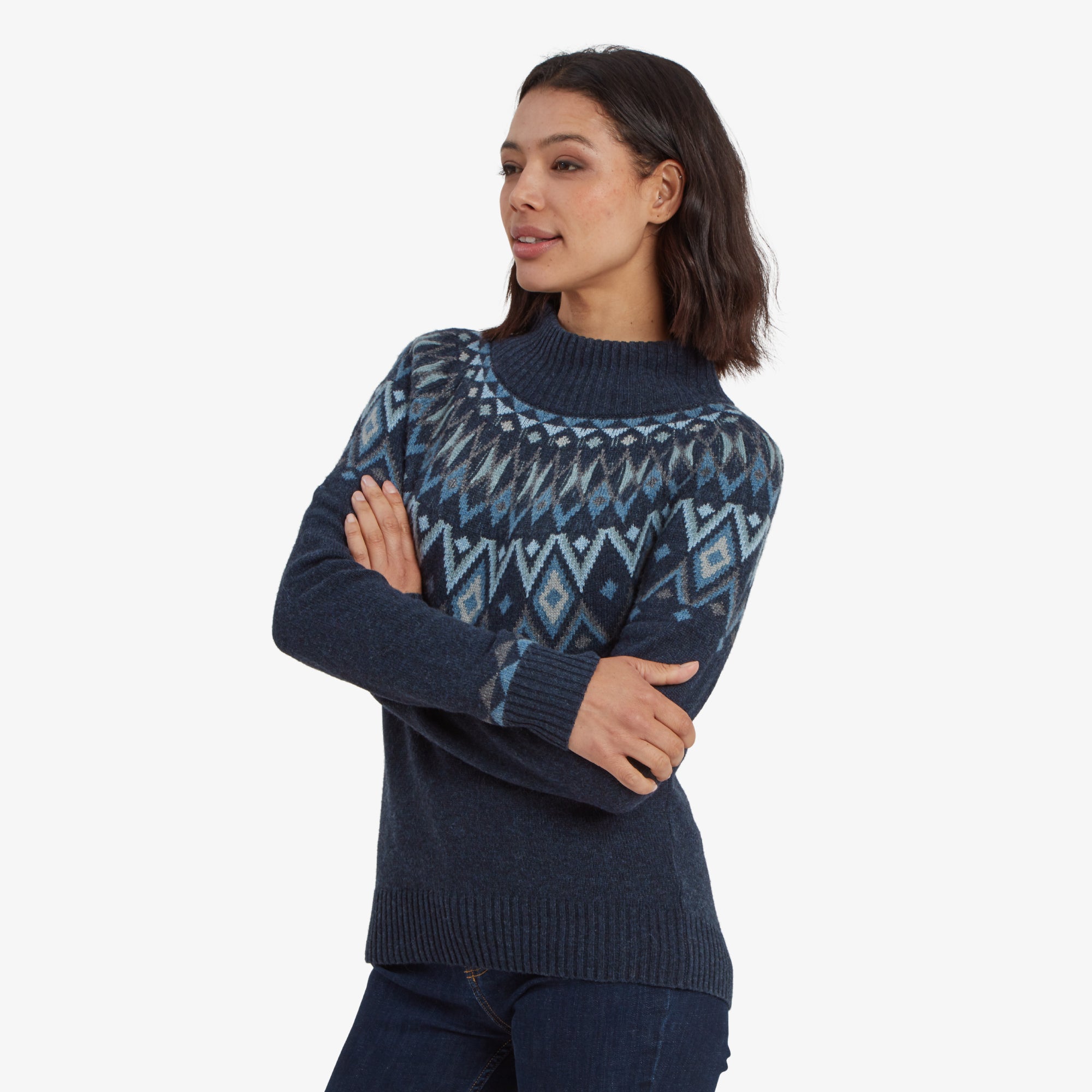 A side-angle view of the woman shows her crossing her arms while wearing the Sherpa Adventure Gear Indu Eco Mock Neck Sweater in Blue. She looks off to the side with a neutral expression, highlighting the fit and ribbed cuffs of the sweater.