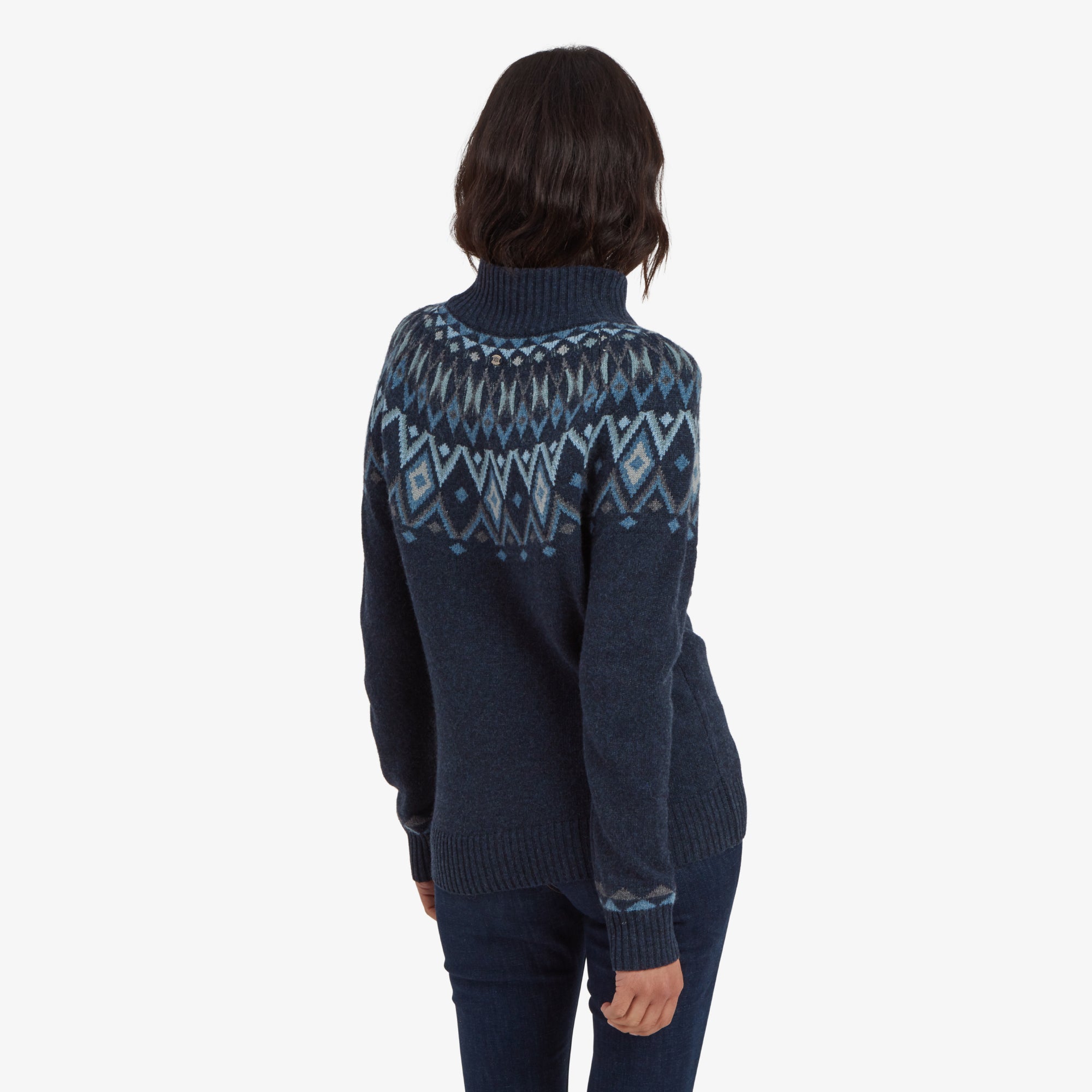 A back view of the Sherpa Adventure Gear Indu Eco Mock Neck Sweater in Blue, showing the continuation of the intricate blue and white geometric pattern across the upper back. The model's short wavy hair frames her face as she slightly turns her head.