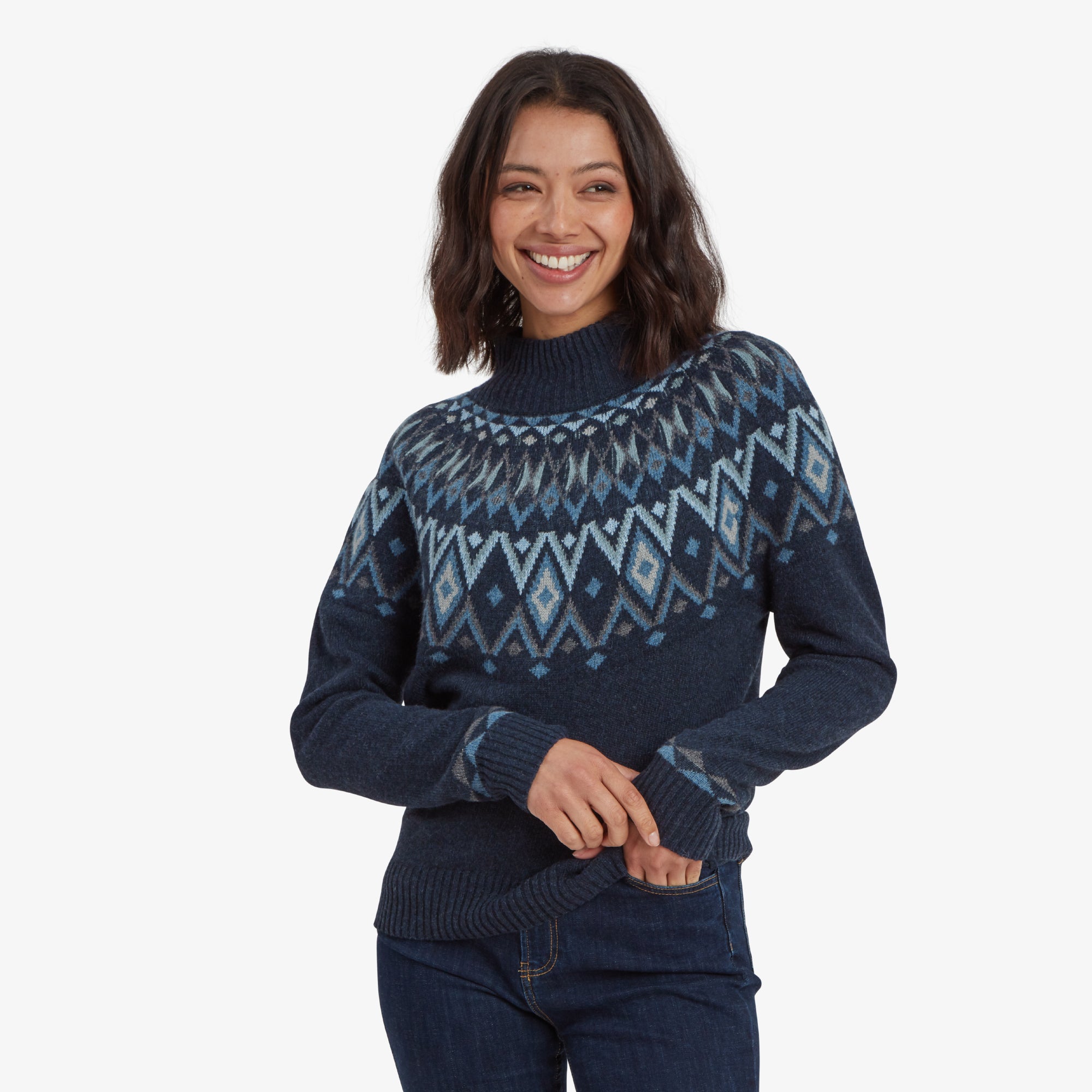 The woman is smiling brightly while holding the hem of the Sherpa Adventure Gear Indu Eco Mock Neck Sweater in Blue with both hands, displaying the snug fit and soft texture of the knit fabric.