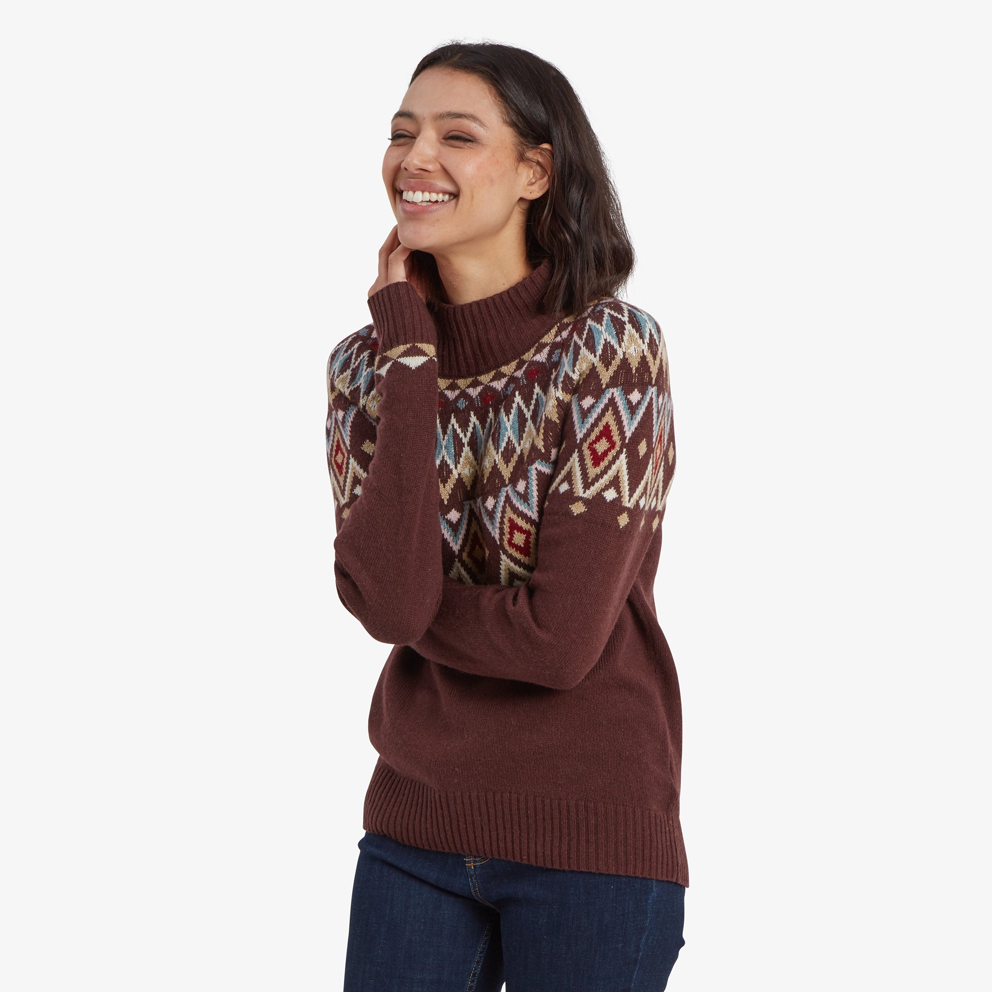 A close-up image of a woman wearing a Sherpa Adventure Gear Indu Eco Mock Neck Sweater in Red with a colorful diamond-patterned yoke. She is smiling and tilting her head slightly, resting her hand on her chin. The sweater has a mock neck design, and the pattern includes hues of blue, cream, and red.