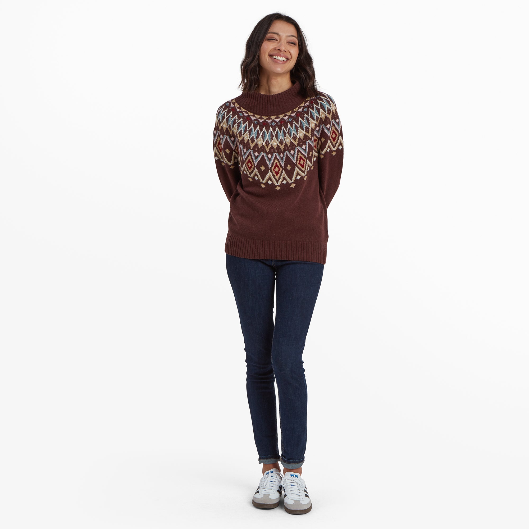 A full-length shot of the woman wearing the same Sherpa Adventure Gear Indu Eco Mock Neck Sweater in Red, paired with dark blue jeans and trainers. She is standing straight with her hands behind her back, smiling brightly. The intricate diamond pattern is prominently displayed across the chest and sleeves.