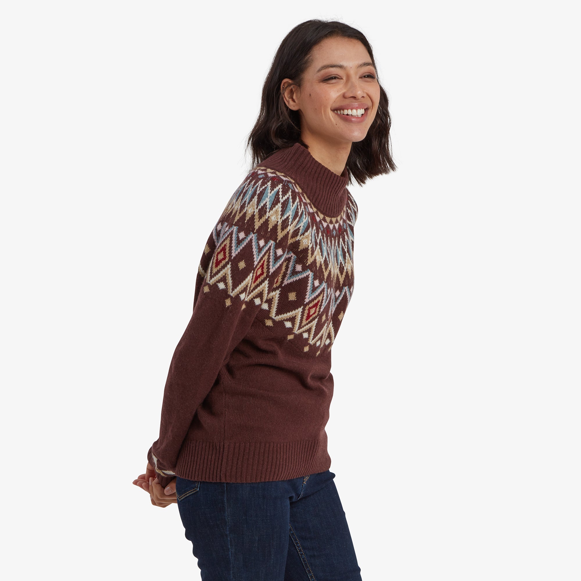 A side angle of the woman wearing the Sherpa Adventure Gear Indu Eco Mock Neck Sweater in Red. She is leaning slightly forward with a cheerful expression, showcasing the diamond yoke design that extends to the upper arms.