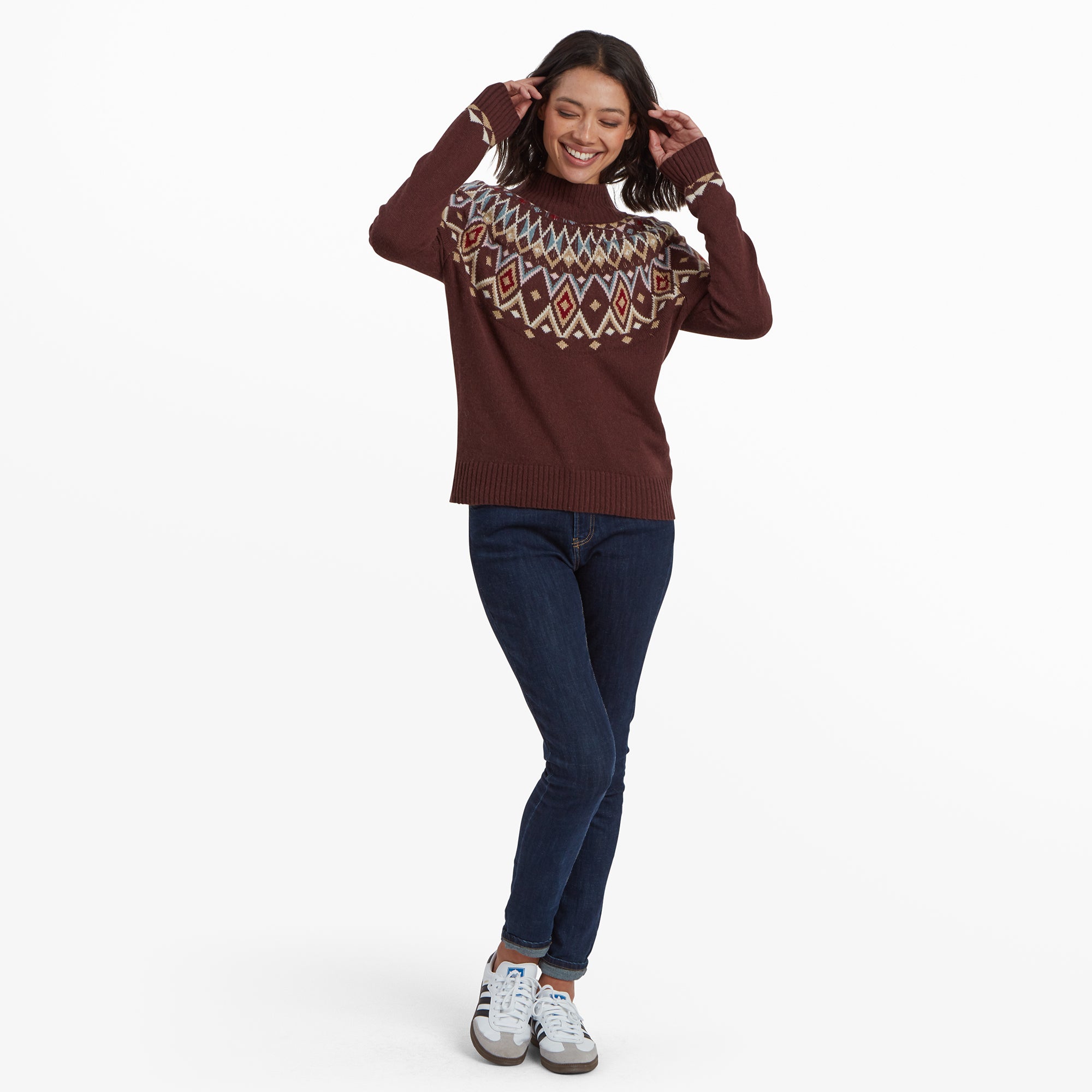 A playful, full-length image of the woman standing with her arms lifted slightly and a wide smile. The Sherpa Adventure Gear Indu Eco Mock Neck Sweater in Red is paired with fitted dark blue jeans and casual trainers, emphasising a cosy yet stylish look.