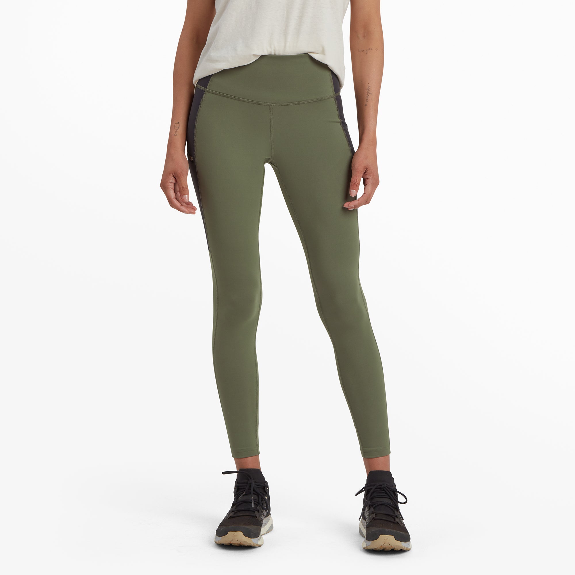 A pair of high-waisted Sherpa Adventure Gear Kalpana Tight in Green with black side panels, modeled by a woman standing straight. She is wearing a light beige t-shirt tucked into the leggings, paired with black trail trainers. The leggings feature a clean, snug fit, highlighting their stretchy and comfortable design.