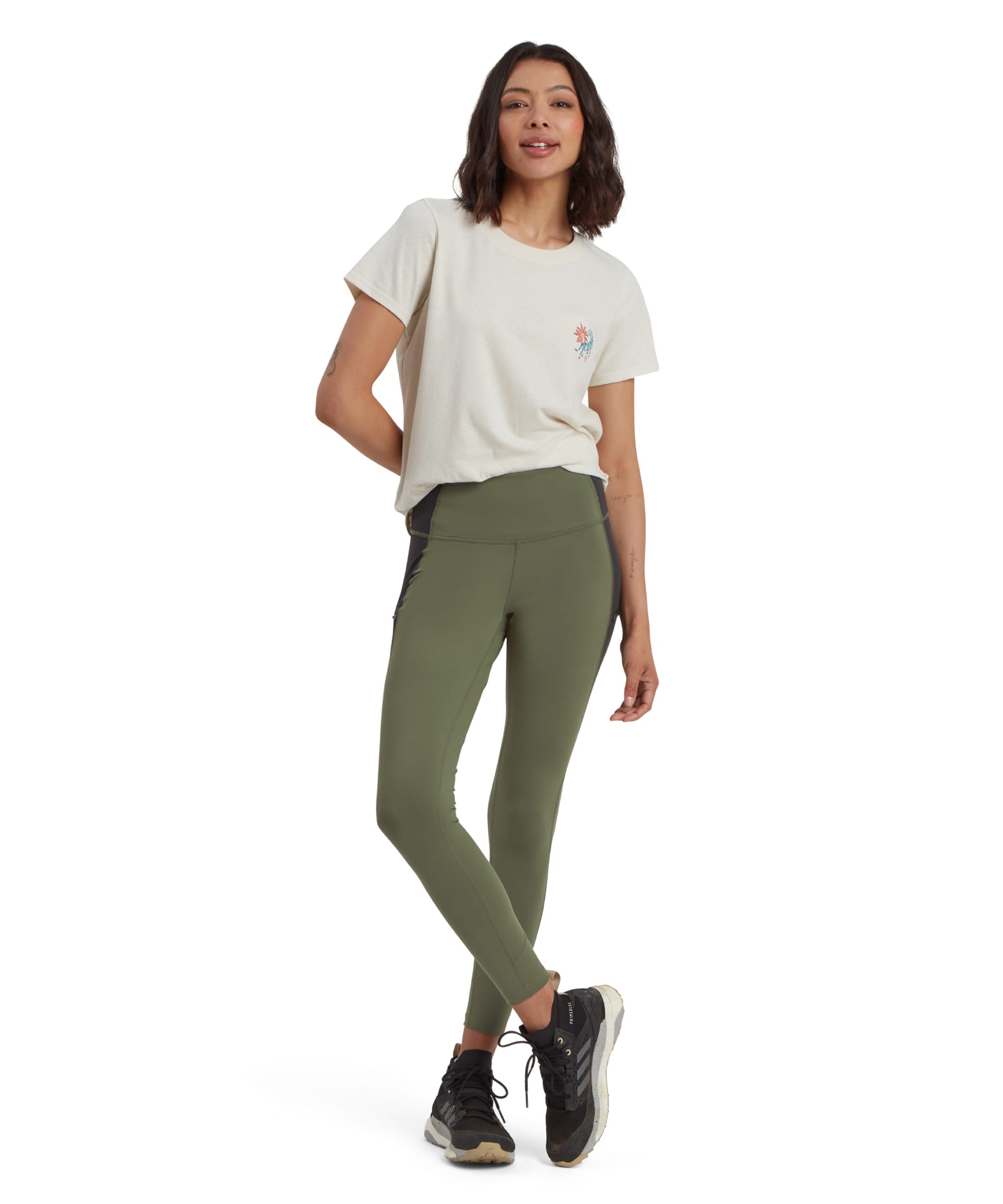 A woman modeling a casual outdoor outfit, consisting of Sherpa Adventure Gear Kalpana Tight in Green with black side panels and a beige graphic t-shirt. She is standing confidently with one leg slightly bent, wearing black trail trainers. The leggings emphasise their activewear style and versatile fit.