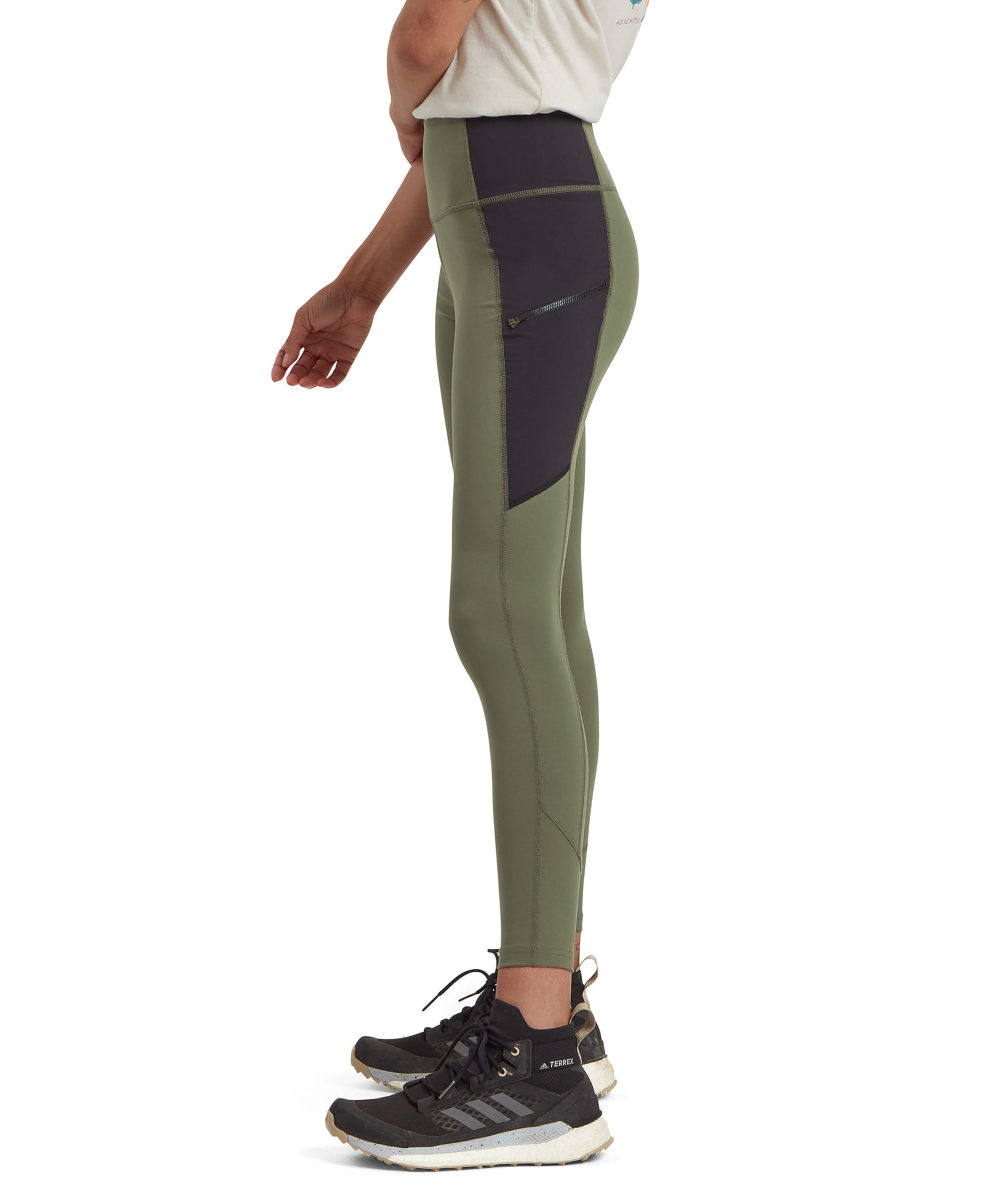 A side profile of Sherpa Adventure Gear Kalpana Tight in Green with black contrast panels extending from the hips down to the calves. The leggings are showcased with a focus on their slim, contouring design and side zipper detail. The model is wearing black trainers and a beige t-shirt.