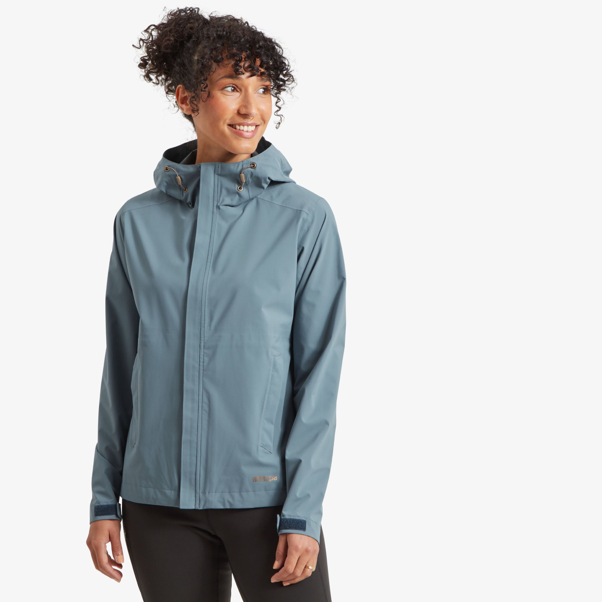 The model is wearing a Sherpa Adventure Gear Nima 2.5-Layer Jacket in Blue with a fully zipped front. The jacket features a clean and modern design with a high neck and an adjustable hood. The fabric appears smooth, with subtle seam detailing for structure. She pairs it with black pants, standing confidently.