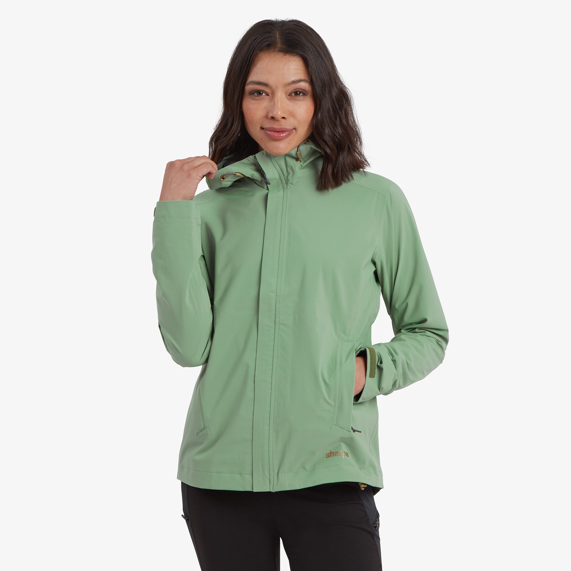 A close-up of a woman wearing a Sherpa Adventure Gear Nima 2.5-Layer Jacket in Green with the hood up, showcasing the adjustable drawstrings and high collar. The jacket has a clean, minimalist design with subtle Sherpa branding.