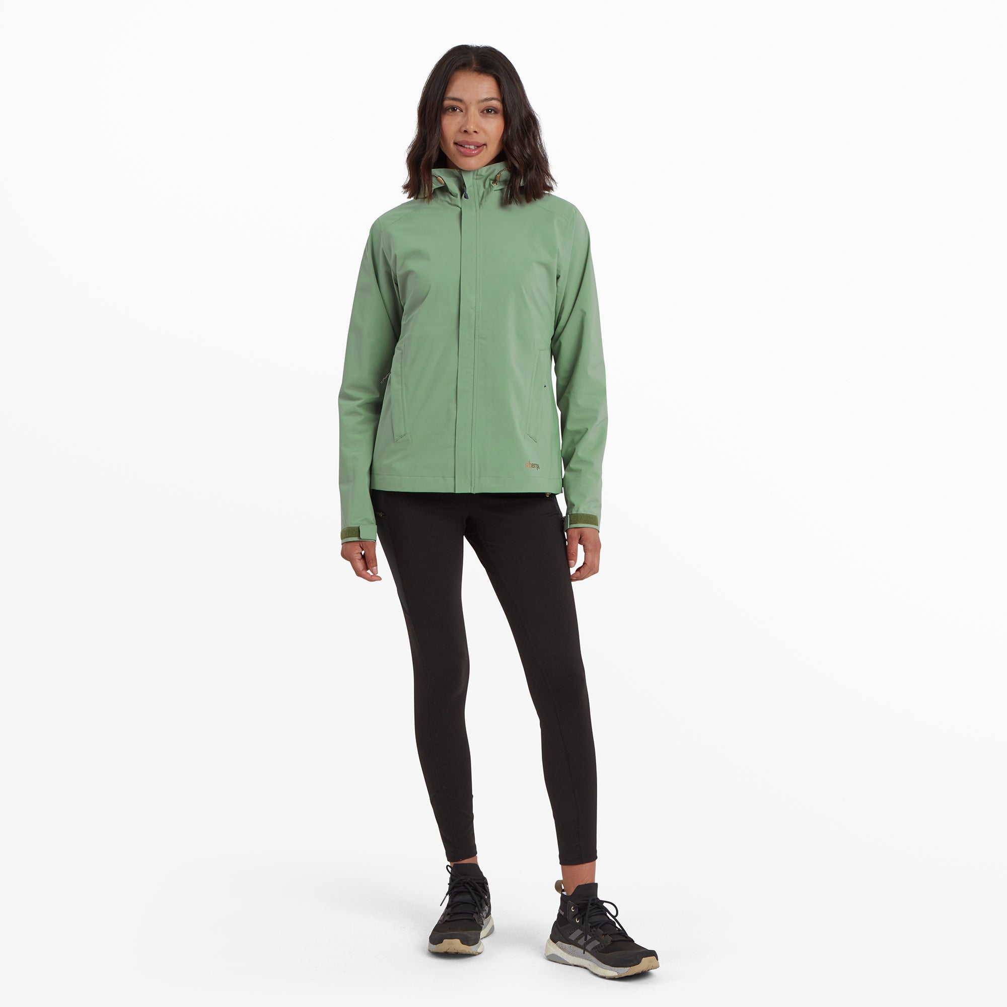 The model stands facing forward, wearing the Sherpa Adventure Gear Nima 2.5-Layer Jacket in Green paired with black leggings and trail shoes. This image highlights the jacket's tailored fit and hip-length design.