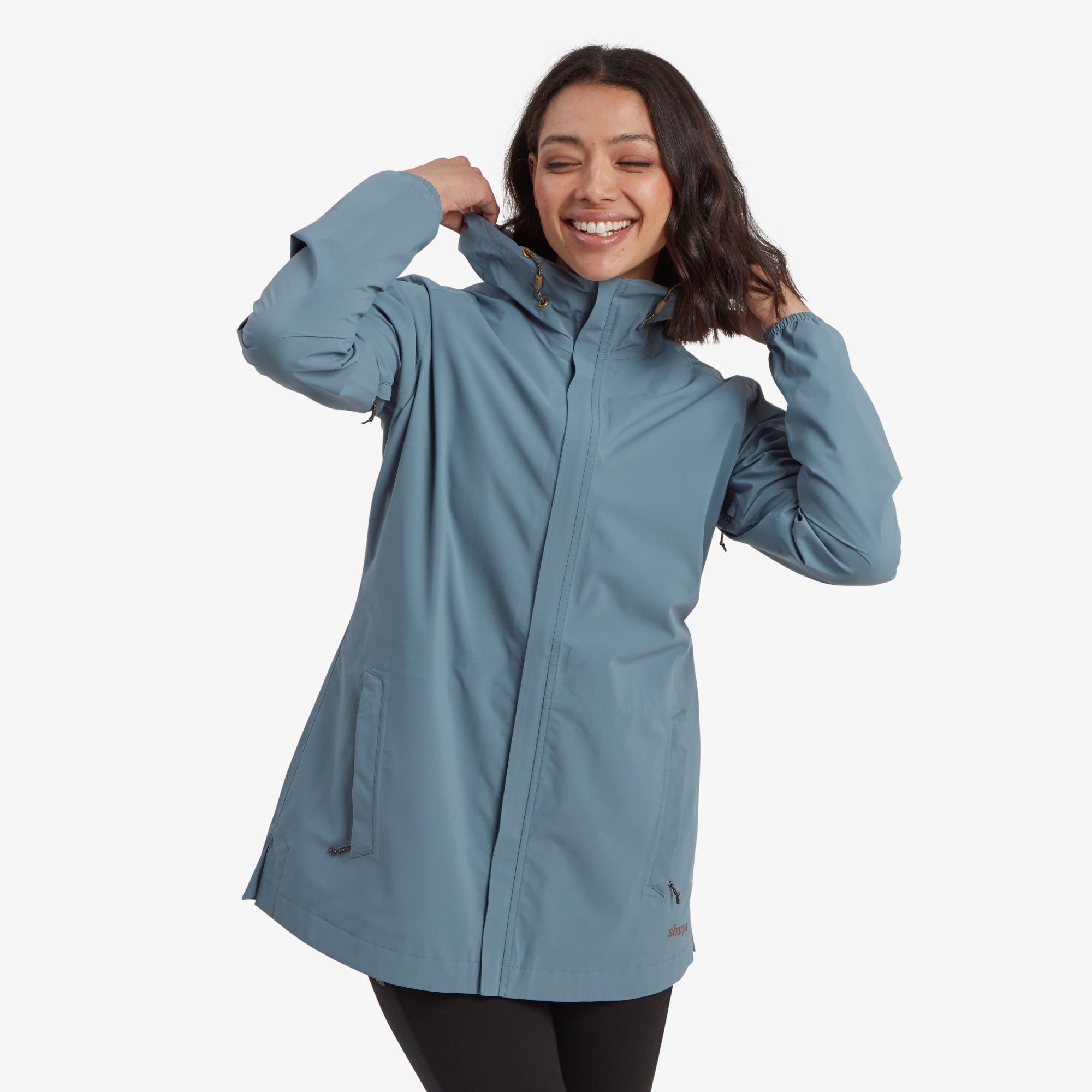 A smiling woman wearing a Sherpa Adventure Gear Nima 2.5-Layer Long Jacket in Blue with a high collar and a hood. She is lifting the collar slightly with her hands, showcasing the jacket's snug and adjustable fit.