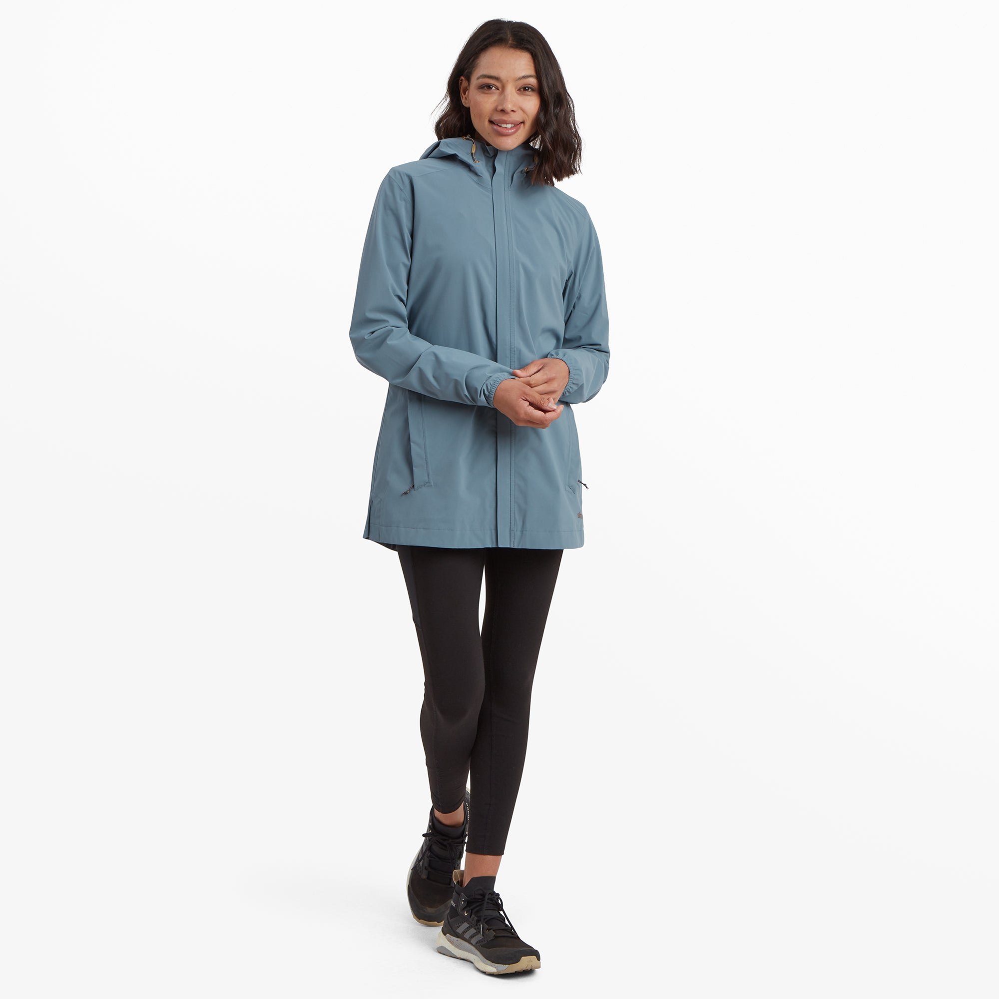 The same woman wearing the Sherpa Adventure Gear Nima 2.5-Layer Long Jacket in Blue, standing upright and fully zipped, displaying its full length. The jacket features sleek side pockets and a clean, minimalist design.