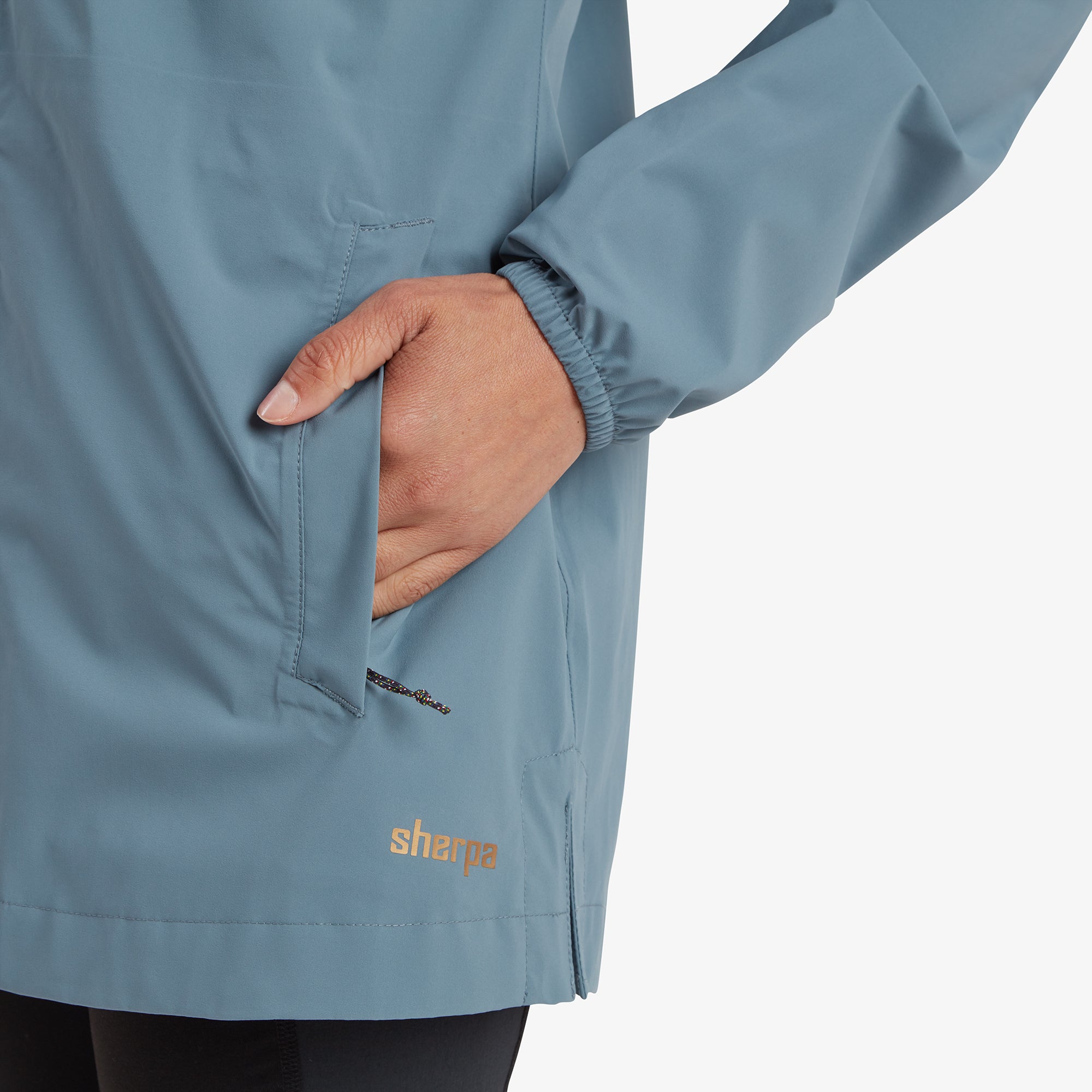 A close-up of the side pocket with the woman’s hand partially inside, highlighting the Sherpa Adventure Gear Nima 2.5-Layer Long Jacket in Blue’s functional pocket design and subtle Sherpa logo near the hemline.