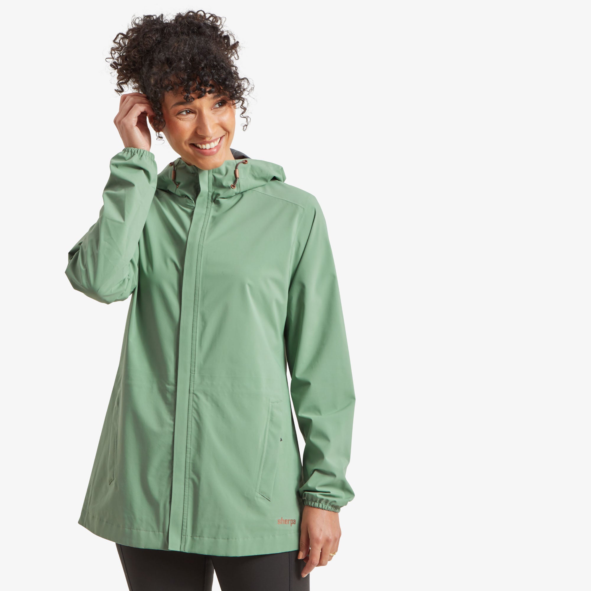 Front view of the jacket. A woman with curly hair is smiling while wearing a Sherpa Adventure Gear Nima 2.5-Layer Long Jacket in Green with a hood. She is adjusting her hair with one hand. The jacket features a front zipper with a storm flap and two zippered pockets. The "Sherpa" logo is printed on the lower left side.