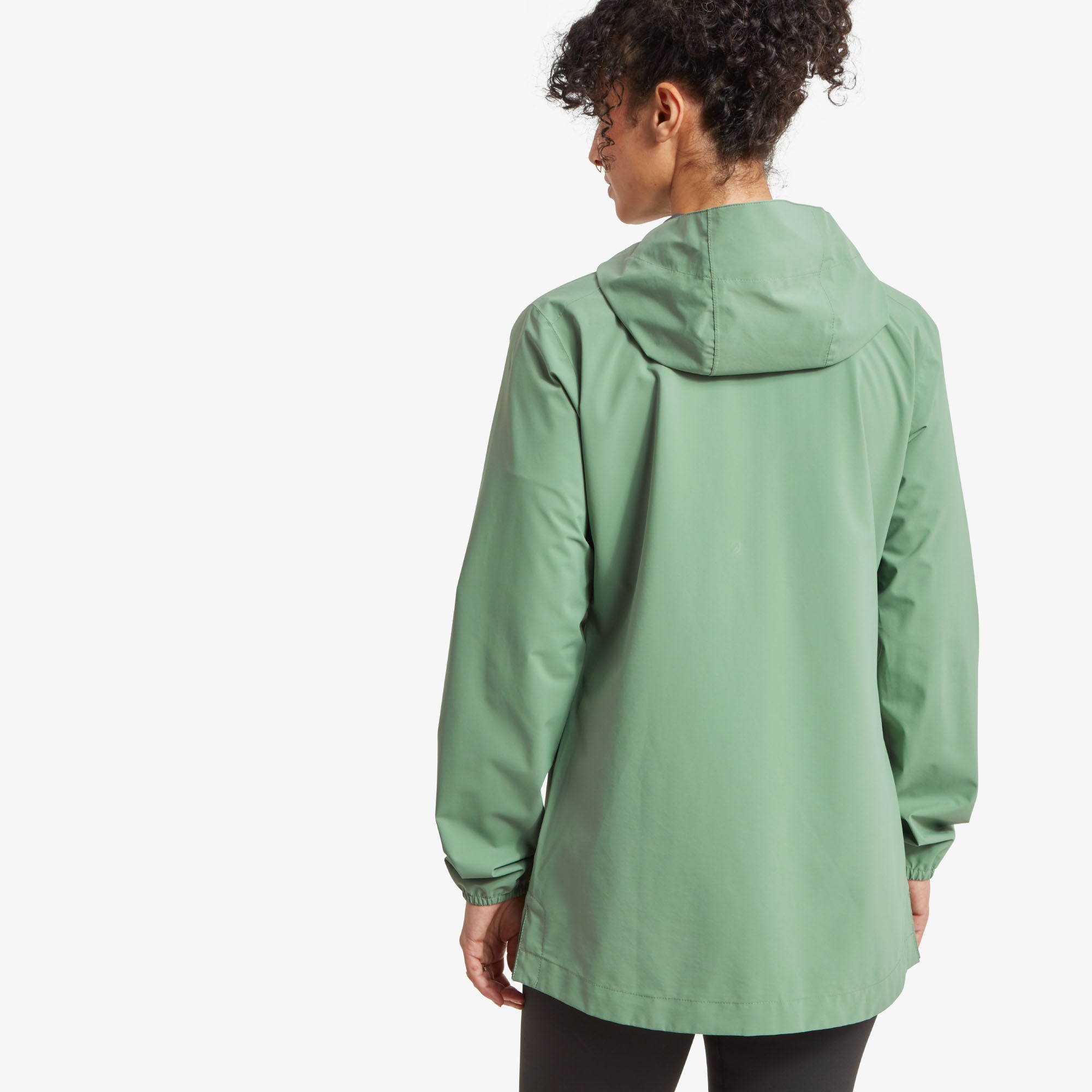 Back view of the jacket. The model is turned away from the camera, showing the back of the Sherpa Adventure Gear Nima 2.5-Layer Long Jacket in Green. The hood is down, revealing its simple design. The sleeves have elastic cuffs, and the hem extends slightly below the hips for extra coverage.