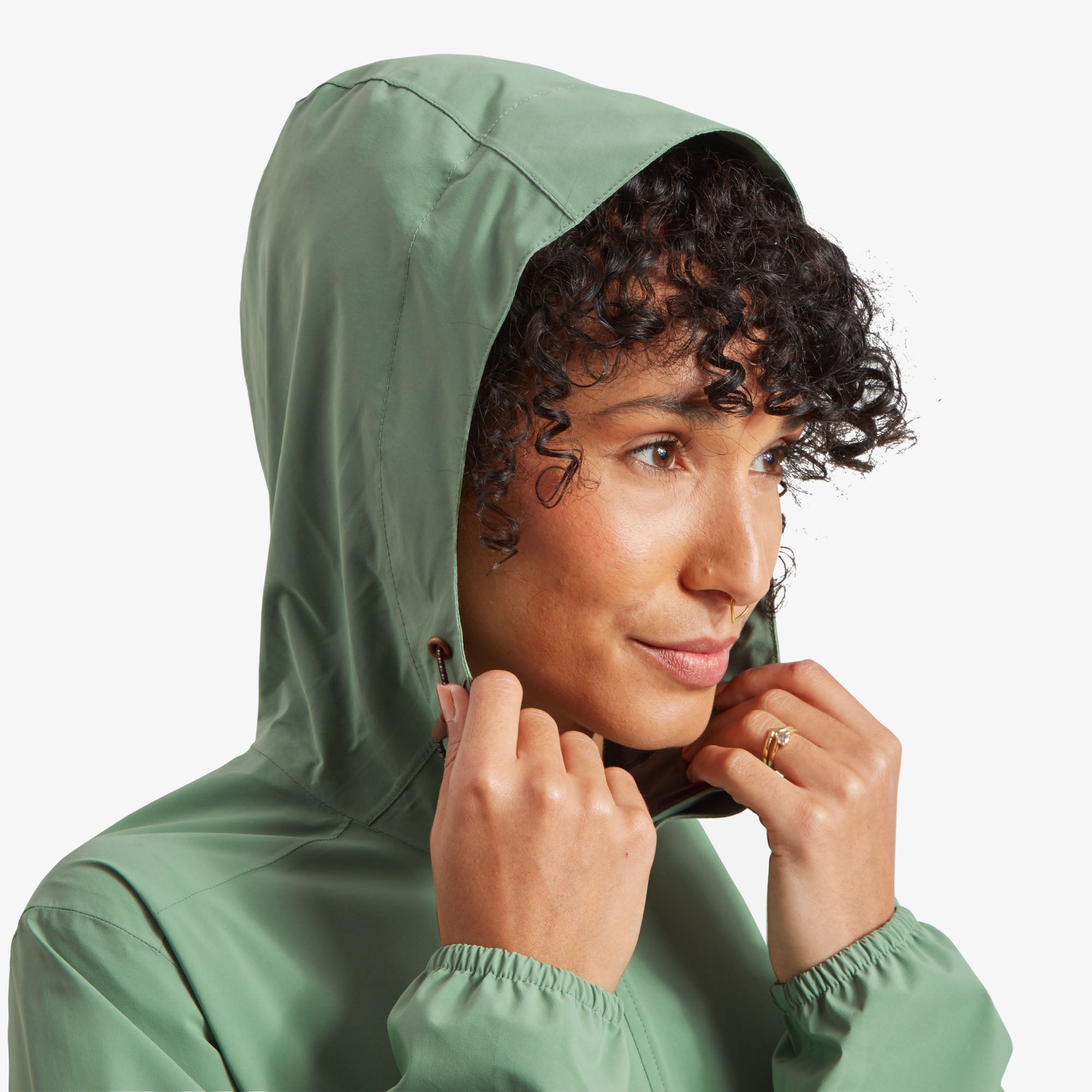 Close-up of the hood in use. A close-up of the model wearing the Sherpa Adventure Gear Nima 2.5-Layer Long Jacket in Green showing the hood, gently pulling the drawstrings. The hood has adjustable cords with brown accents and metal stoppers. Her face is partially visible with a soft smile.