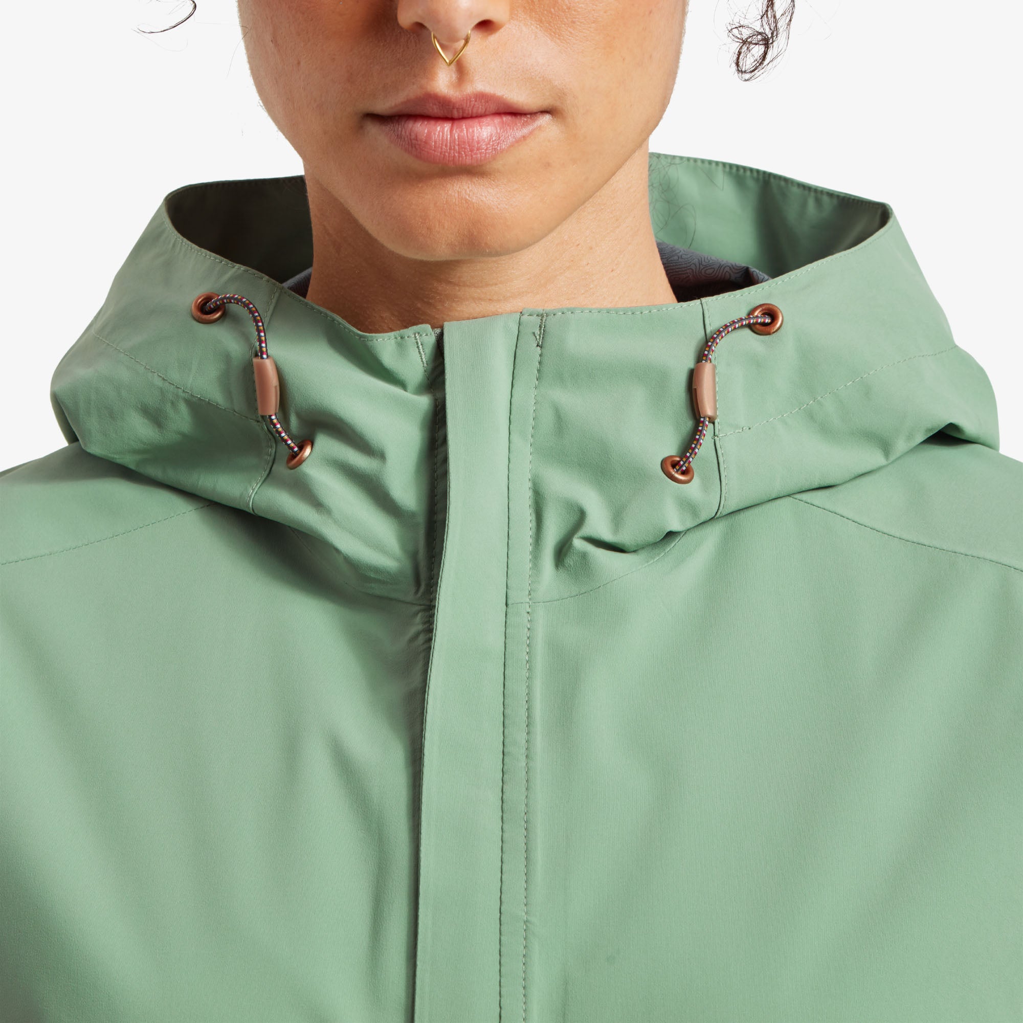 Close-up of the high collar and hood drawstrings. A tight shot of the model’s face and the upper part of the Sherpa Adventure Gear Nima 2.5-Layer Long Jacket in Green, highlighting the high collar and hood drawstrings. The light green fabric is smooth and waterproof, with brown cord adjusters providing a stylish contrast.