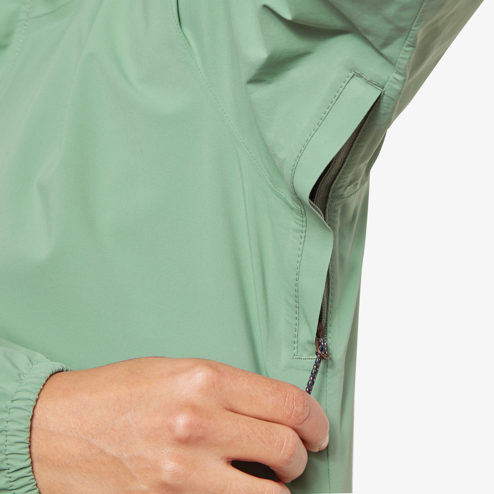 Close-up of underarm ventilation zipper. The model's hand is unzipping a hidden underarm ventilation opening, revealing a breathable mesh layer beneath on the Sherpa Adventure Gear Nima 2.5-Layer Long Jacket in Green. The zipper pull is attached to a black and multicolor cord for ease of use.