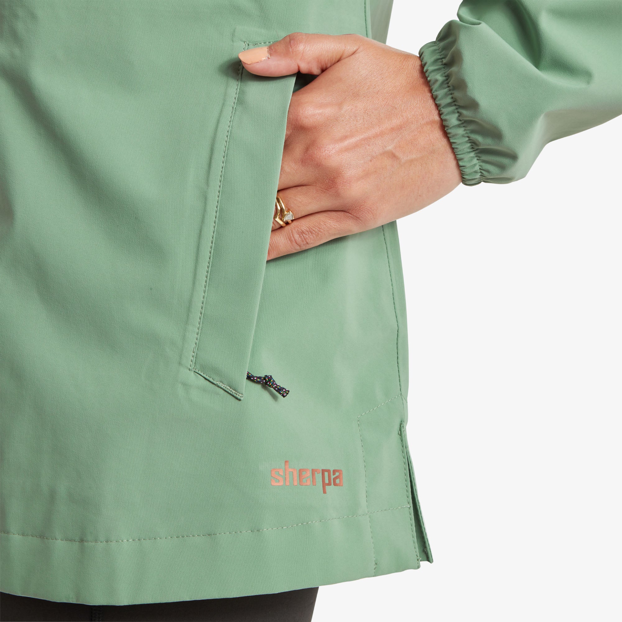 Close-up of a zippered pocket and branding. The model is placing her hand inside a zippered pocket on the lower left side of the Sherpa Adventure Gear Nima 2.5-Layer Long Jacket in Green. The "Sherpa" logo is printed in coral near the pocket. The pocket has a discreet water-resistant zipper.