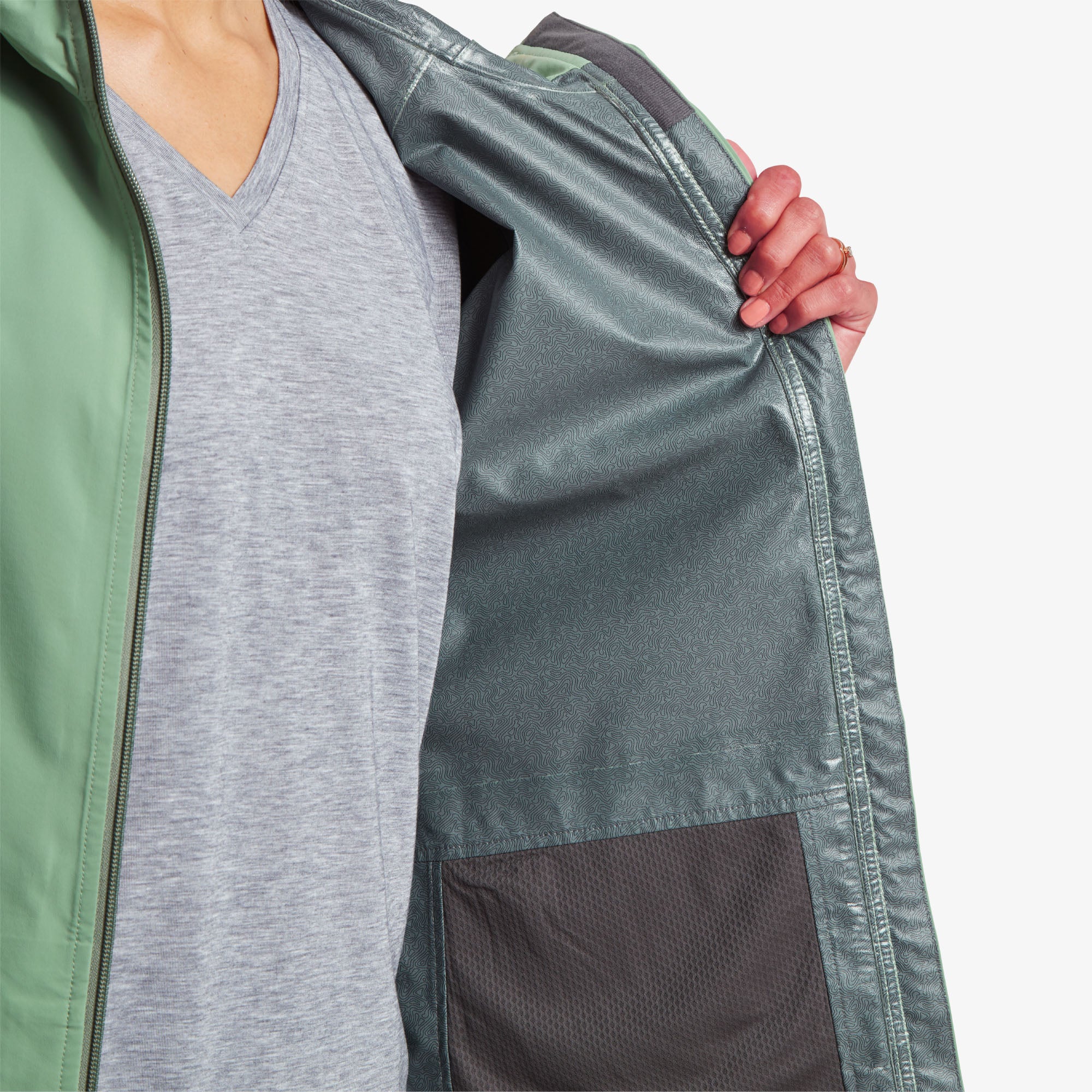 Inside lining of the jacket. The model is holding the Sherpa Adventure Gear Nima 2.5-Layer Long Jacket in Green open, showing its interior lining. The lining is dark teal with a subtle textured pattern and mesh pockets. The waterproof outer shell is visible along the edges.