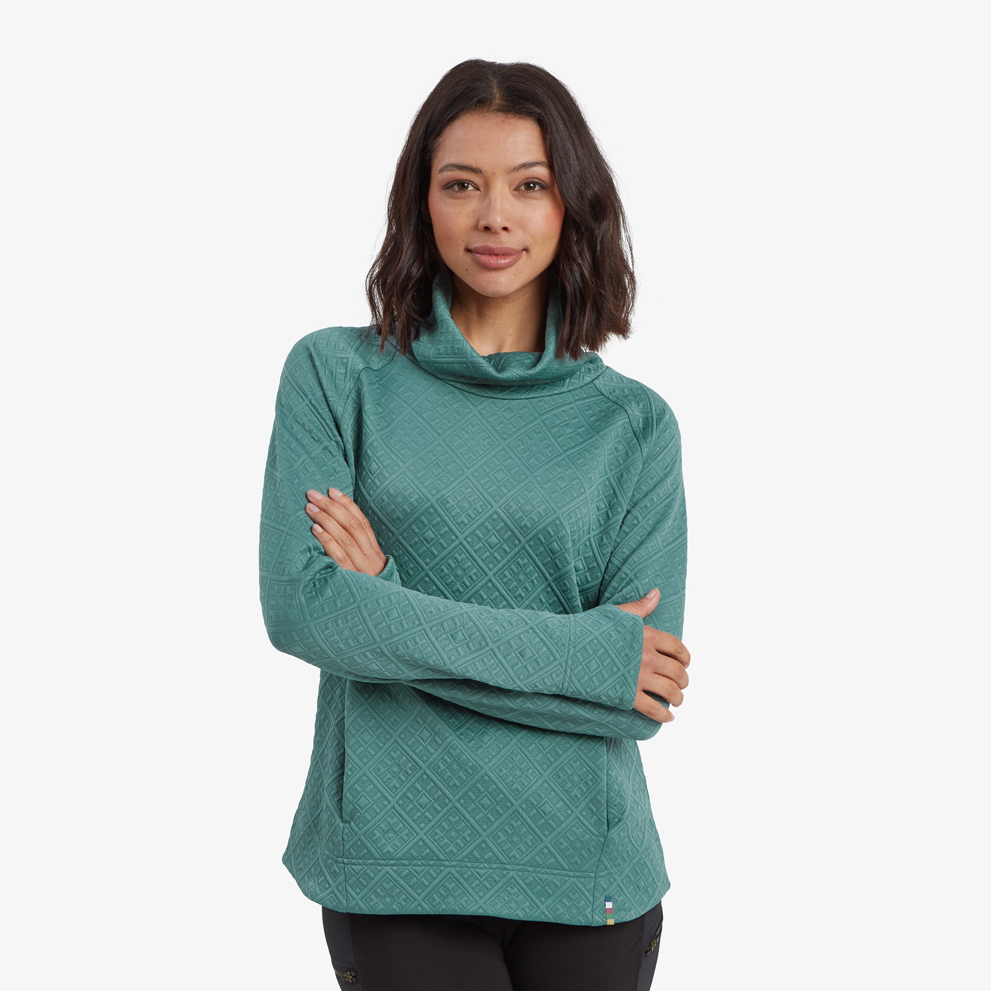 A model wearing a Sherpa Adventure Gear Nyano Pullover in Blue. The pullover features a relaxed fit, quilted diamond pattern texture, and a cozy cowl neck. The long sleeves include thumb holes, and the front pocket is subtly detailed with colorful stitching accents near the hem. The model has her arms crossed and is standing against a plain white background, emphasising the details of the garment.