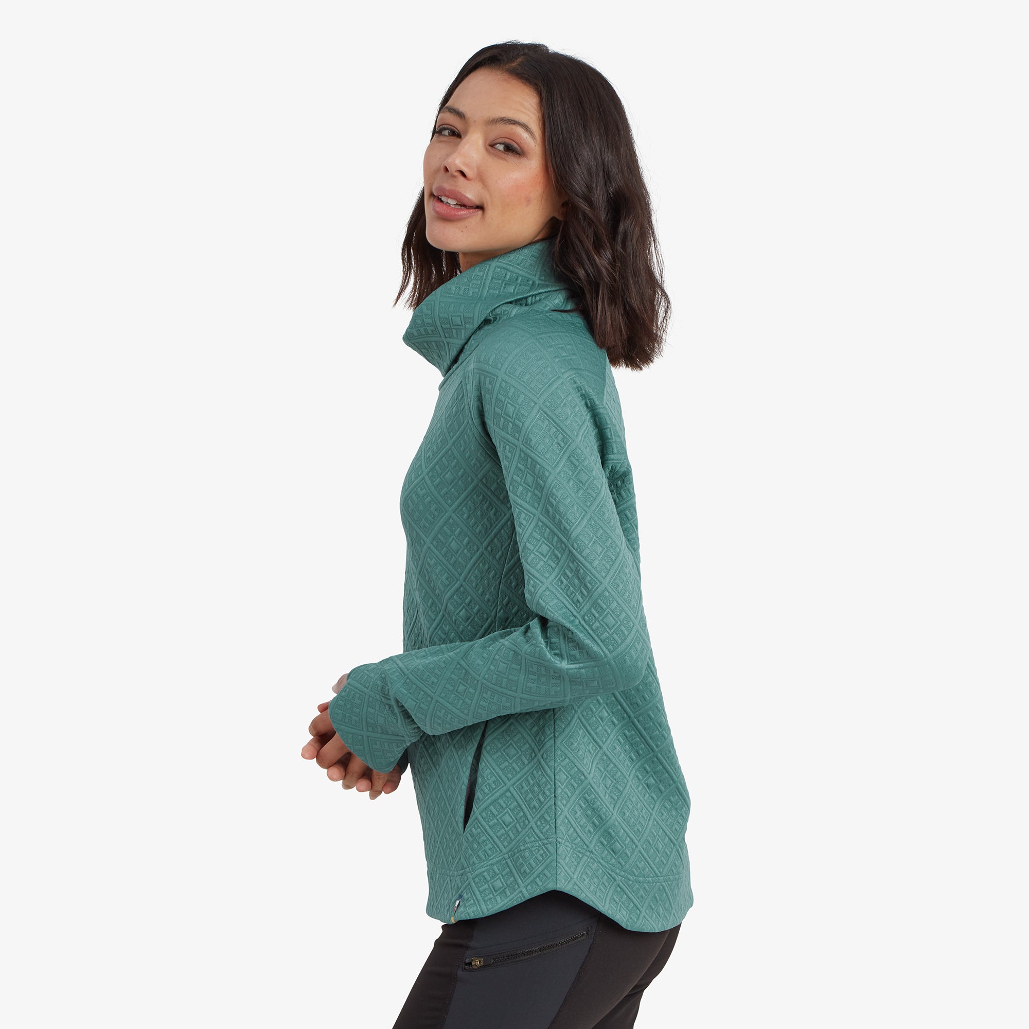 The model is shown in profile, showcasing the side silhouette of the Sherpa Adventure Gear Nyano Pullover in Blue. The slightly curved hemline at the back provides a flattering fit. The quilted diamond texture of the fabric is visible, adding dimension to the design.