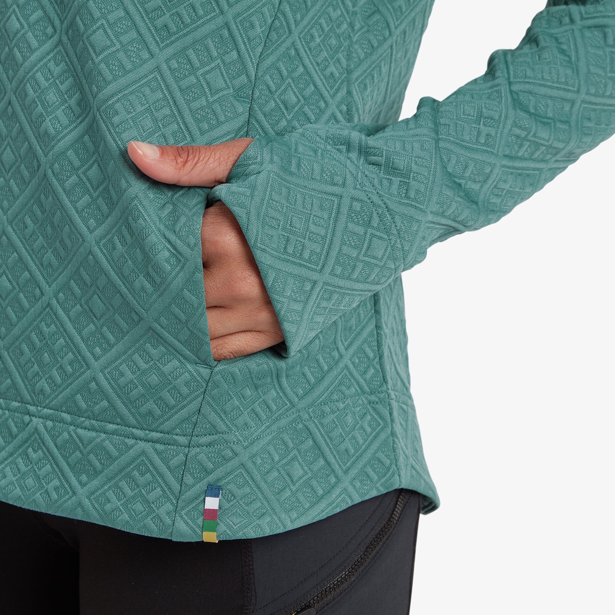 A close-up of the front pocket of the Nyano Pullover, showing its practical design and subtle colorful stitching detail near the hem. The model's hand is resting near the pocket, illustrating its functionality and clean design.