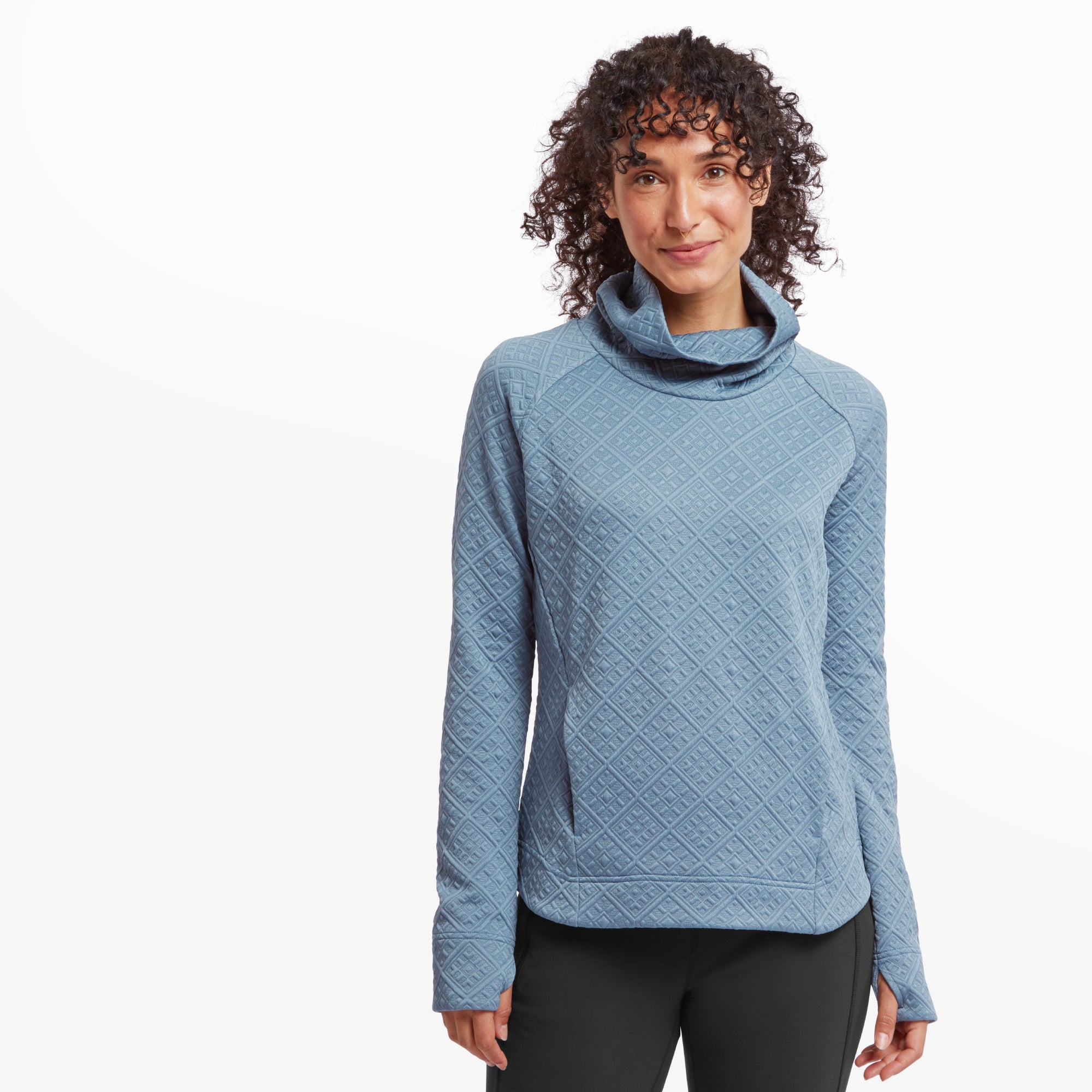 A woman is wearing a Sherpa Adventure Gear Nyano Pullover in Blue with a cowl neck. The quilted texture is highlighted, creating a cozy and structured look. She is standing against a white background with her hands resting at her sides, smiling softly.