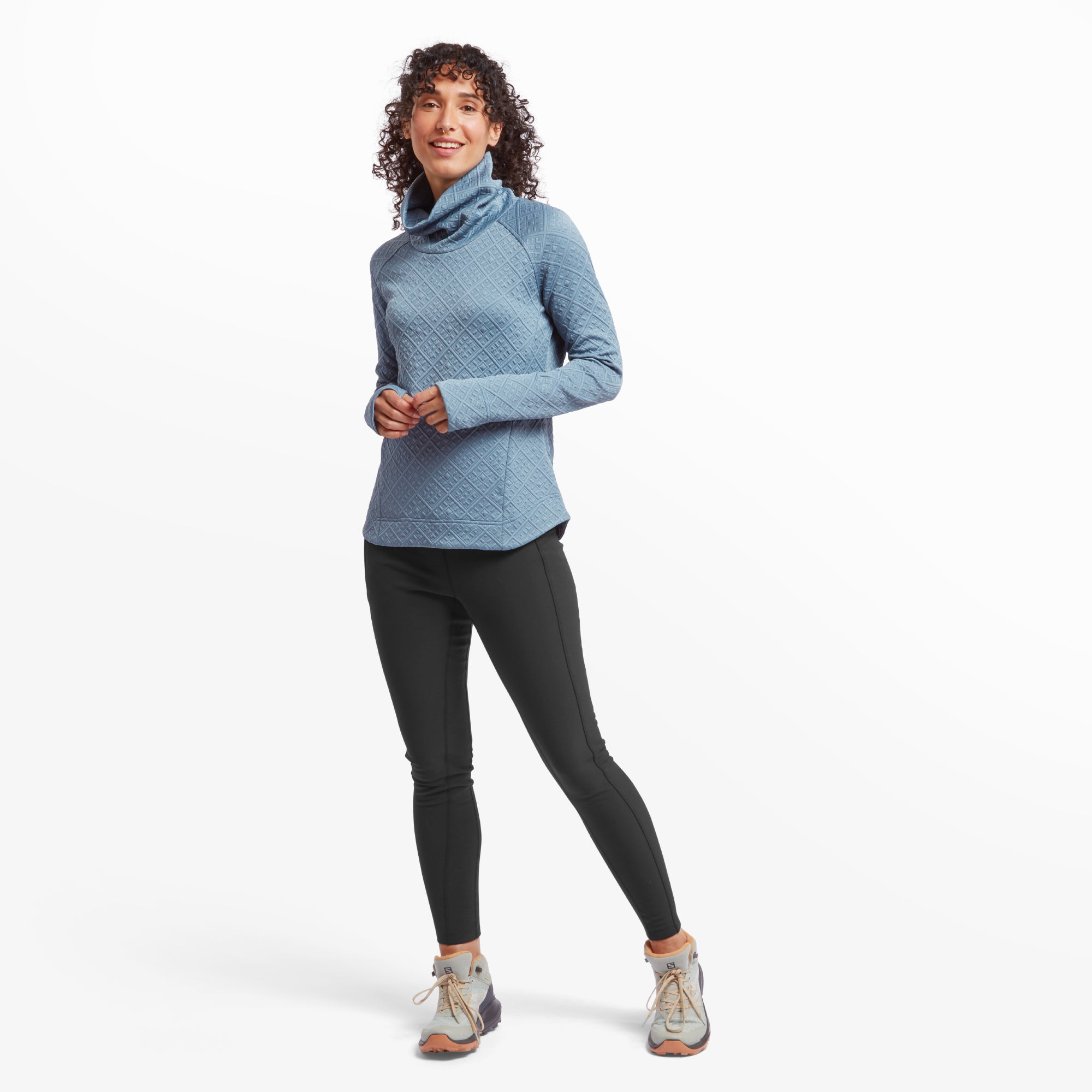 The same woman models the Sherpa Adventure Gear Nyano Pullover in Blue, paired with black leggings and hiking shoes. She poses in a relaxed stance, showcasing the pullover's slightly curved hem and its casual, comfortable fit.